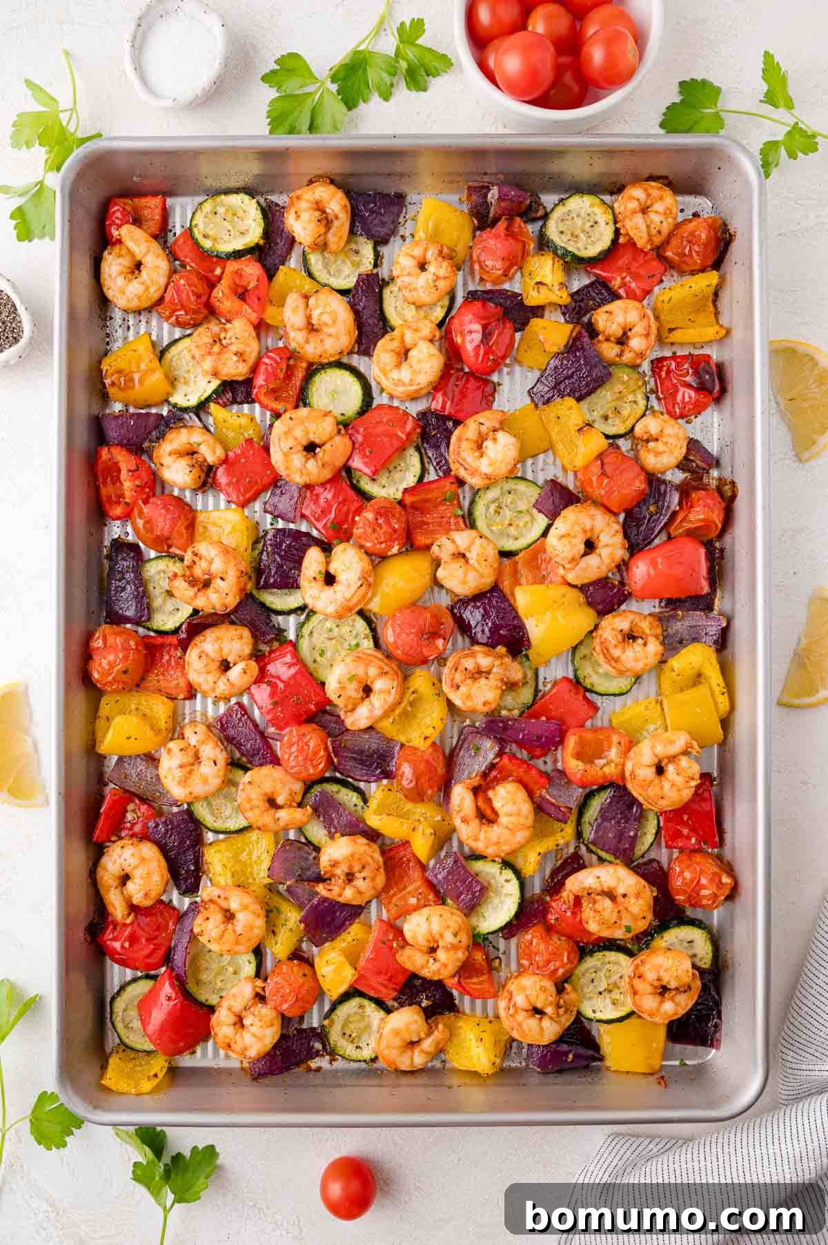 A baking sheet with roasted shrimp, zucchini, yellow and red bell peppers, red onions, and cherry tomatoes, arranged in a colorful pattern. Parsley and lemon wedges are nearby.