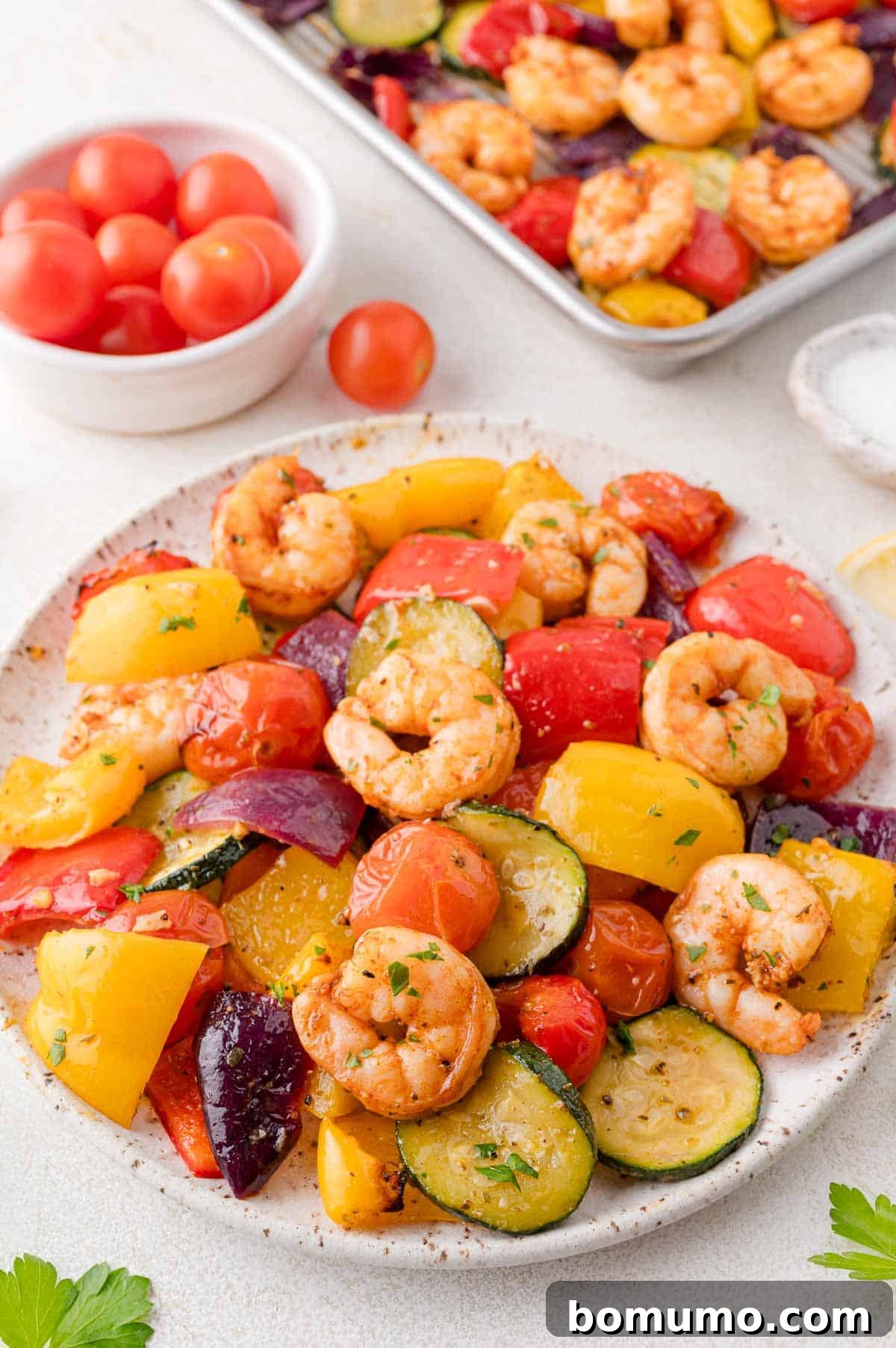 Plate of shrimp with colorful vegetables like yellow peppers, zucchini, and cherry tomatoes, garnished with herbs. In the background, a bowl of cherry tomatoes and a tray with more shrimp and veggies.