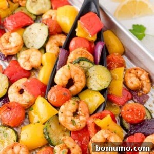 Pan of roasted shrimp with a mix of colorful vegetables including zucchini, bell peppers, cherry tomatoes, and red onions served with a black serving spatula.