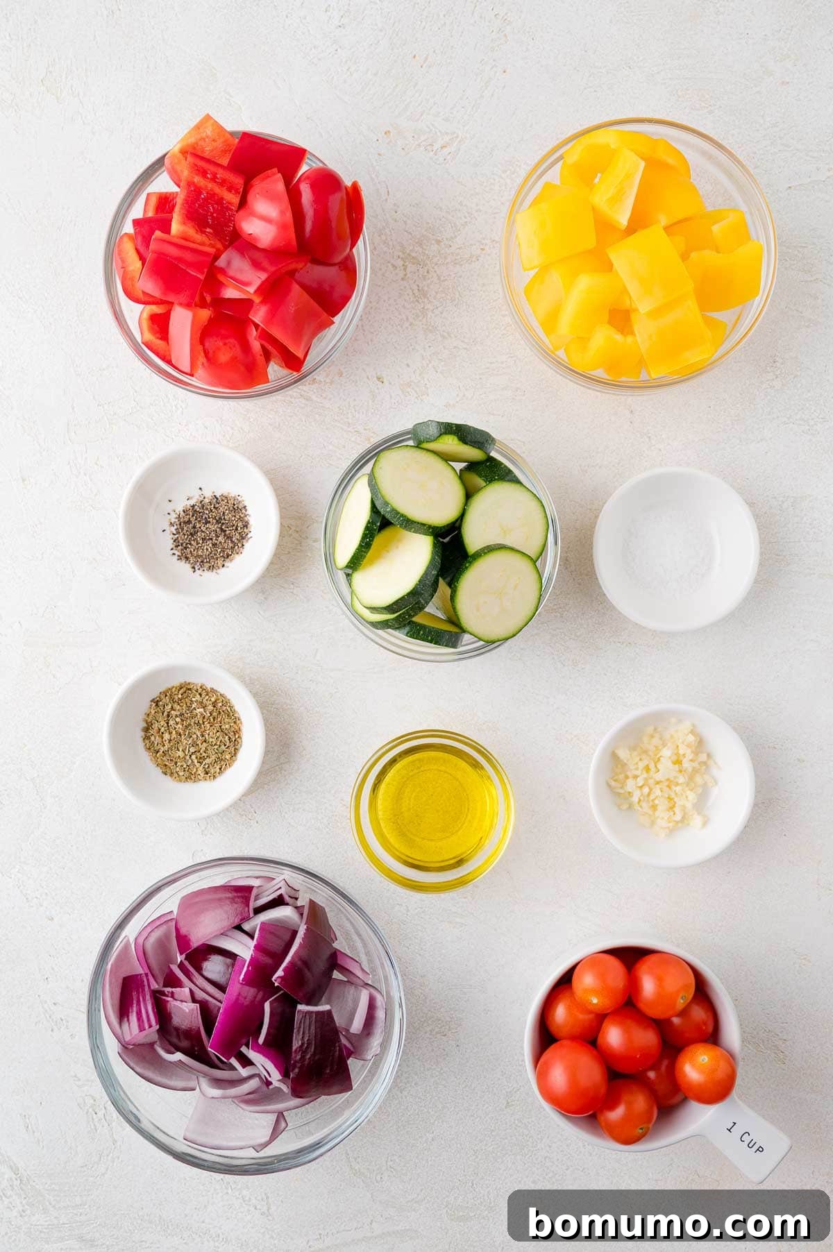 Bowls with diced red and yellow bell peppers, sliced zucchini, cherry tomatoes, chopped red onion, olive oil, minced garlic, and small bowls of salt and pepper on a white surface.
