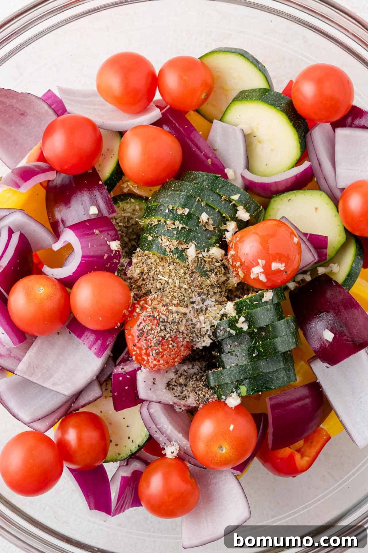 A bowl of raw vegetables including cherry tomatoes, zucchini slices, red onion, and bell peppers, topped with seasoning.