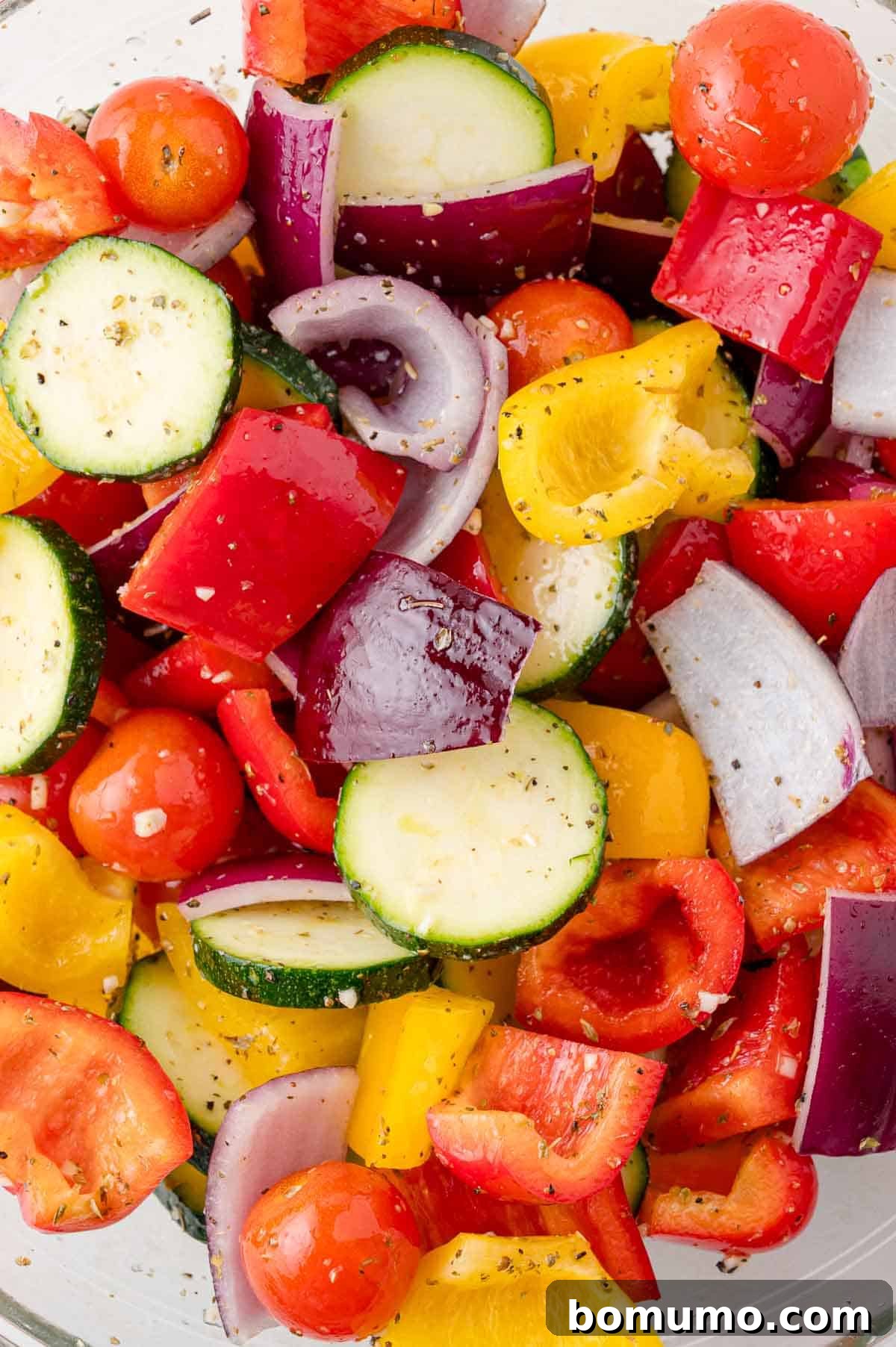 A colorful assortment of chopped vegetables, including zucchini, red onions, cherry tomatoes, and red, yellow, and orange bell peppers, seasoned with herbs and spices.