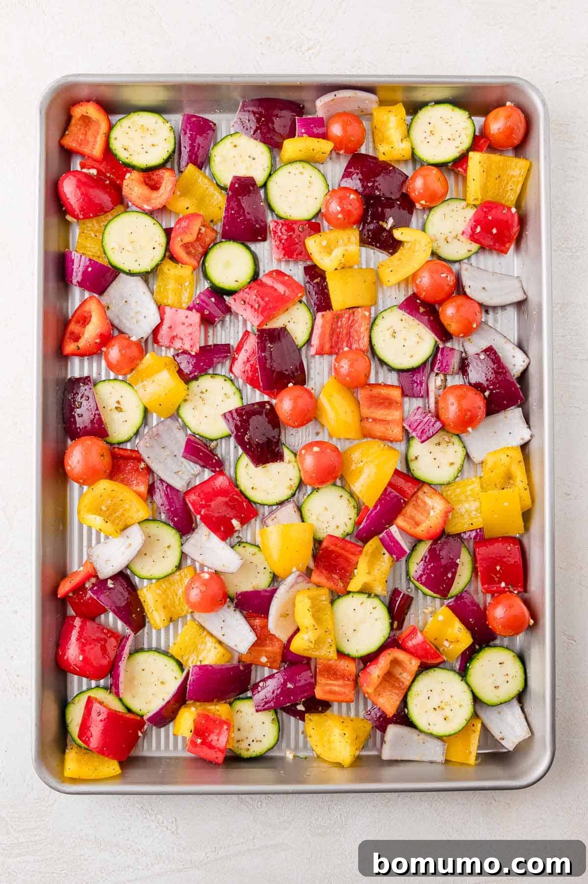 A baking tray filled with chopped vegetables including zucchini, red onion, cherry tomatoes, red and yellow bell peppers, seasoned with herbs.