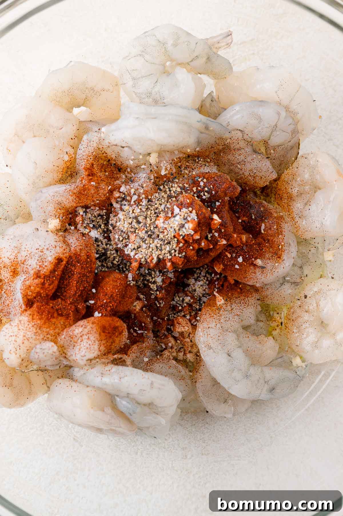 Raw shrimp in a bowl seasoned with paprika, black pepper, and salt.