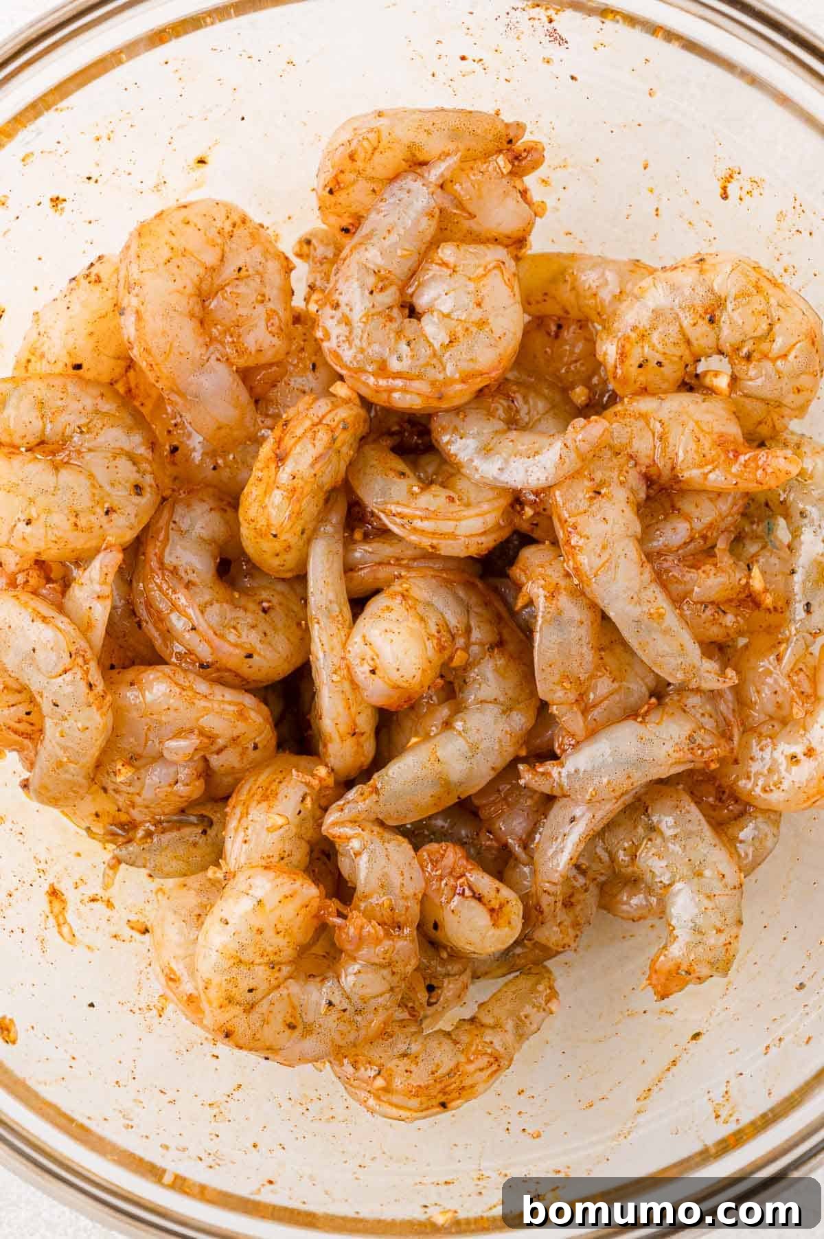 A glass bowl filled with raw shrimp seasoned with a reddish spice mix.