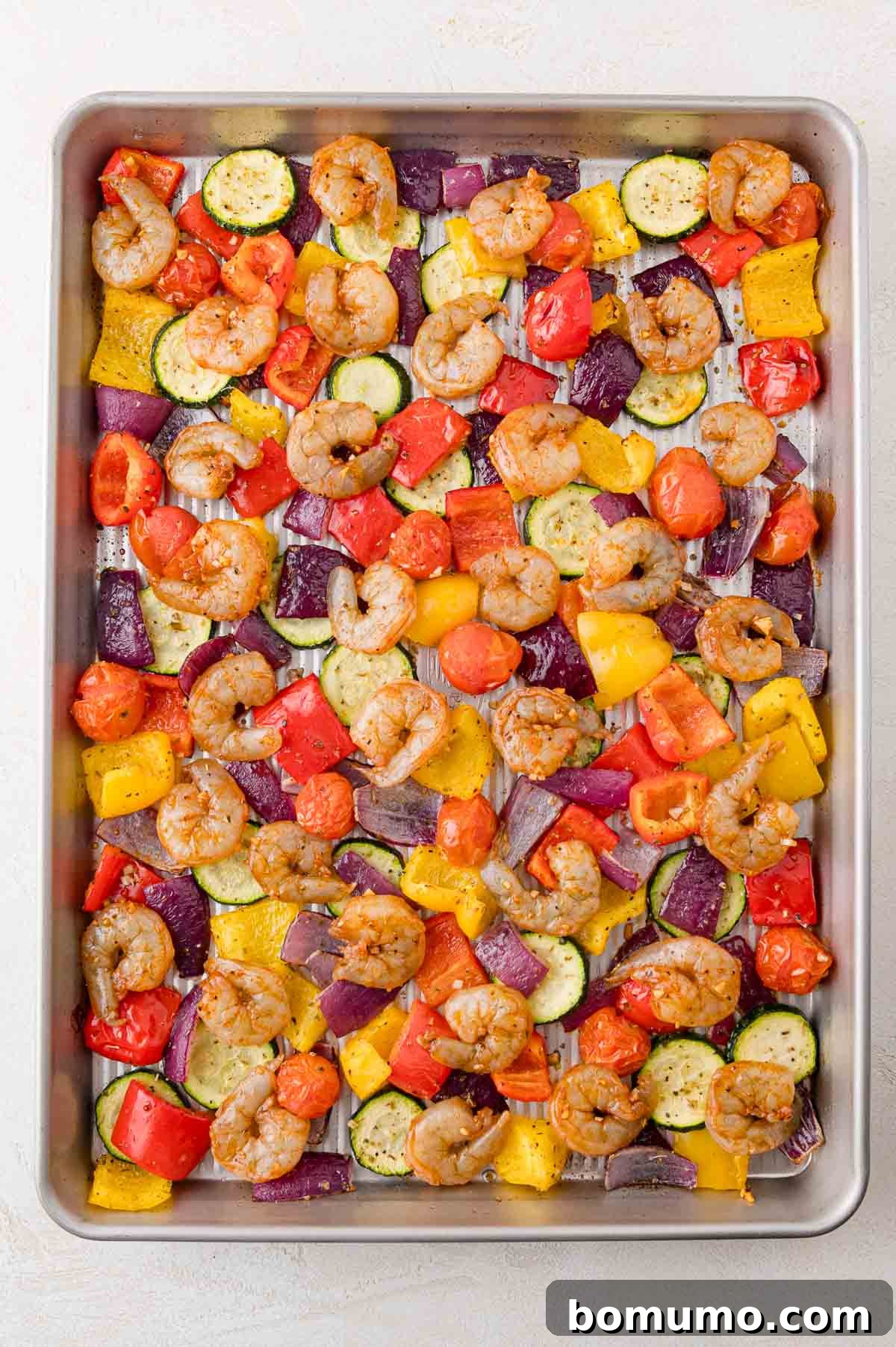 A baking sheet with raw shrimp, sliced zucchini, diced bell peppers, halved cherry tomatoes, and chopped red onions, seasoned and ready to bake.