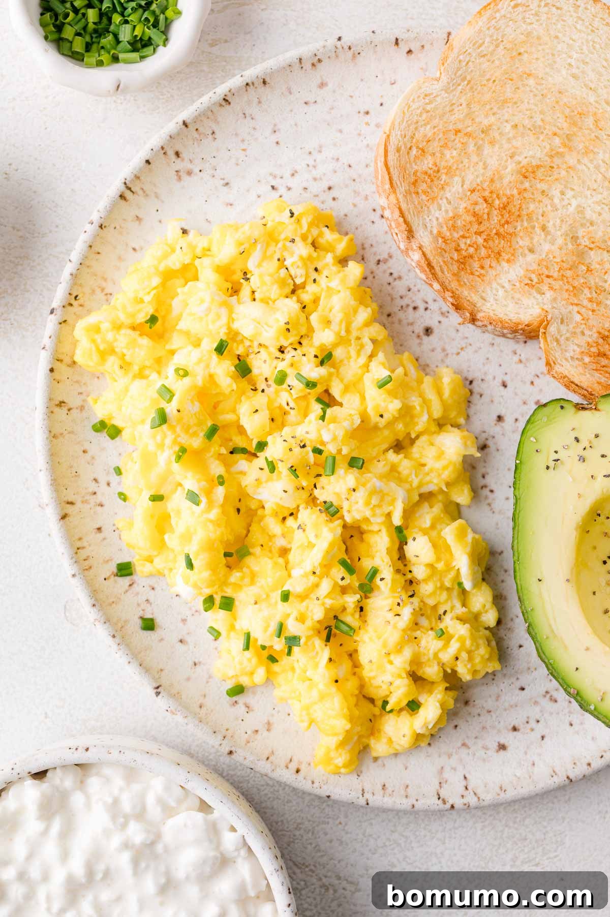 A plate featuring fluffy cottage cheese scrambled eggs, accompanied by a slice of golden toast, and half an avocado, elegantly garnished with freshly chopped chives.