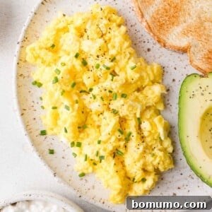 Cottage cheese scrambled eggs garnished with chives, served with sliced avocado and toasted bread on a speckled plate.