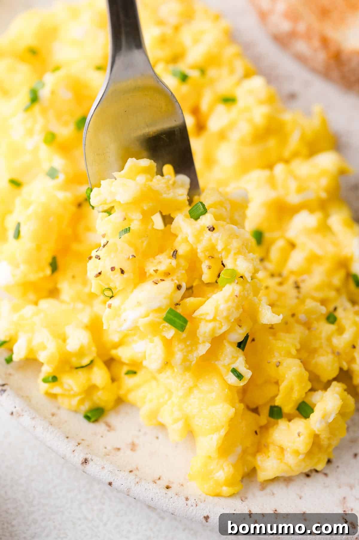 A fork holding a perfectly cooked portion of scrambled eggs, delicately garnished with chopped chives and a sprinkle of freshly ground black pepper, presented on a stylish speckled plate.