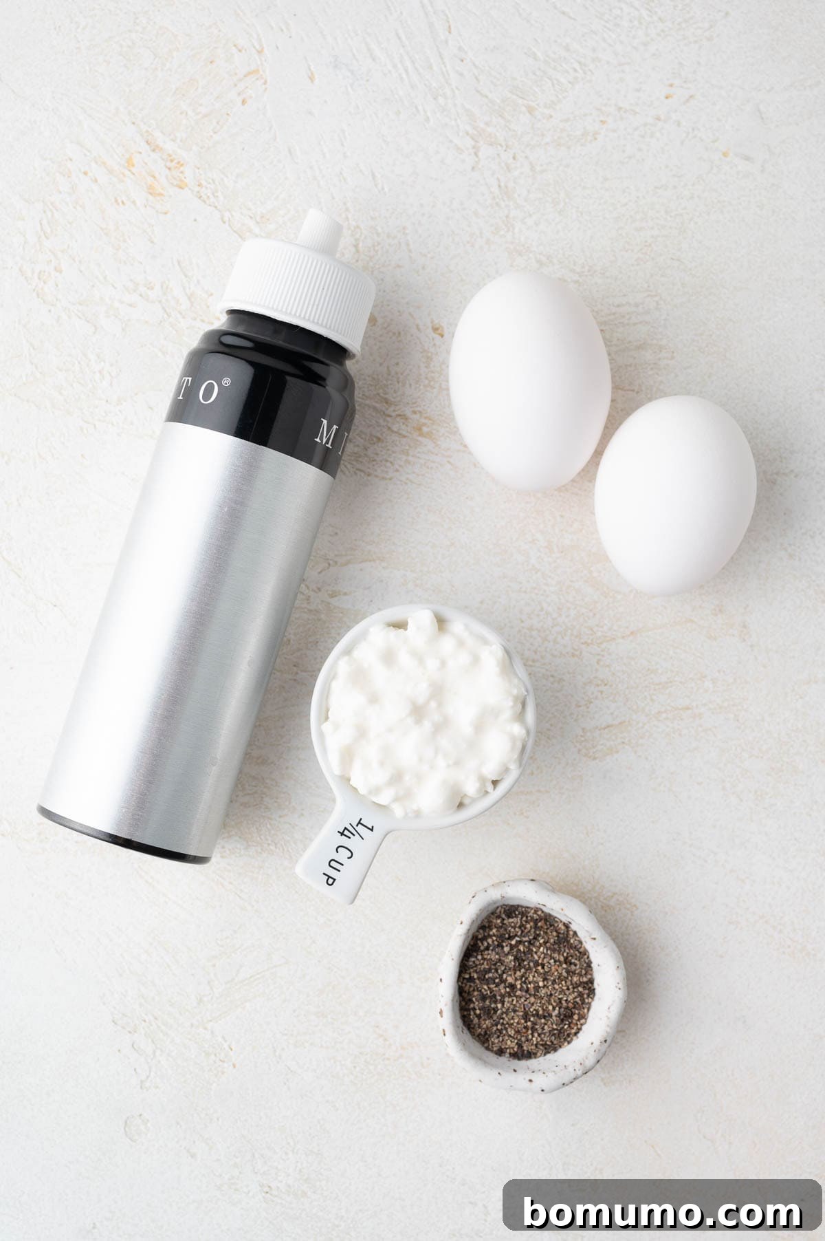 A bottle of cooking oil spray, two fresh eggs, a quarter cup of cottage cheese, and a small bowl of ground black pepper are neatly arranged on a bright, minimalist surface, showcasing the primary ingredients.