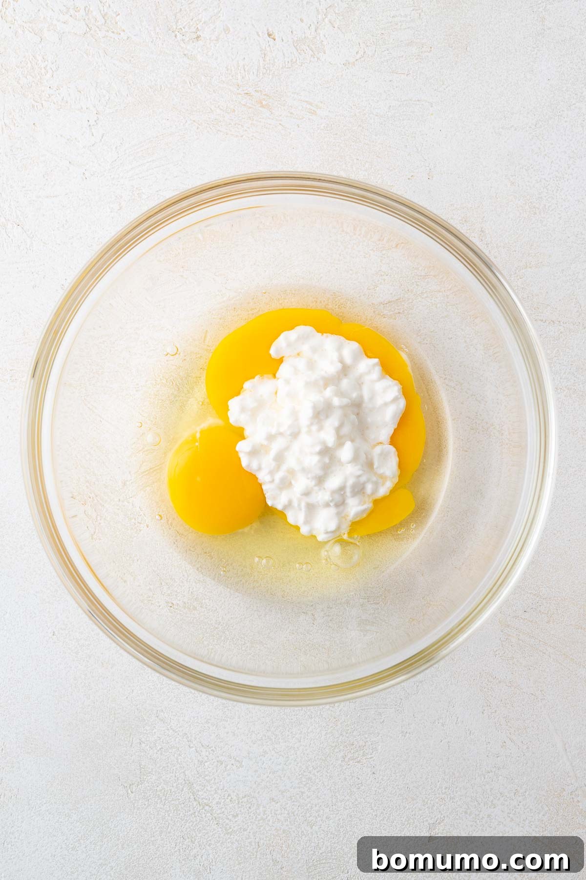 A clear glass bowl containing two raw eggs and a generous portion of white small curd cottage cheese rests on a clean, white surface, ready for mixing.