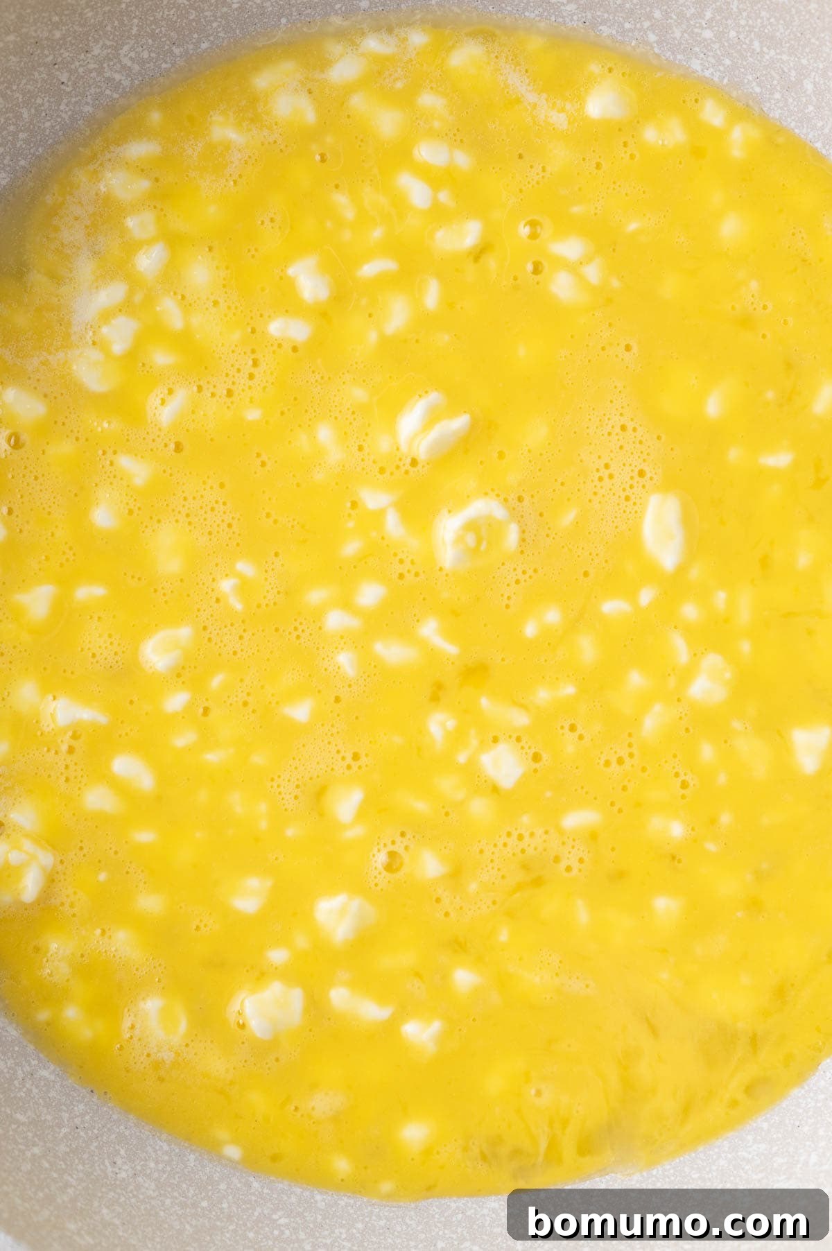 A close-up shot of the mixed beaten eggs and small curd cottage cheese in a bowl, displaying a vibrant yellow hue interspersed with distinct white curds, ready for cooking.