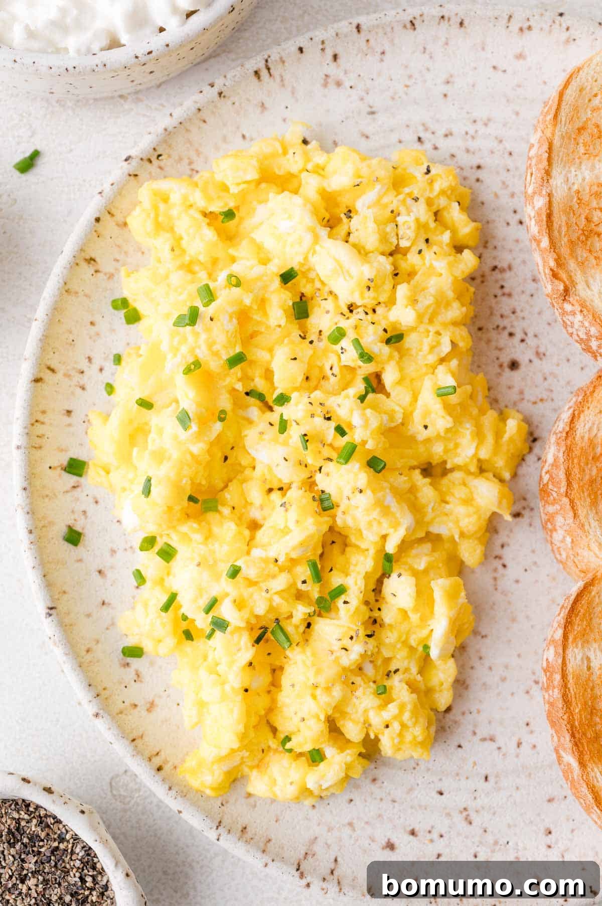 A close-up of perfectly cooked cottage cheese scrambled eggs, garnished with vibrant chopped chives, presented on a speckled plate alongside two slices of golden toasted bread.