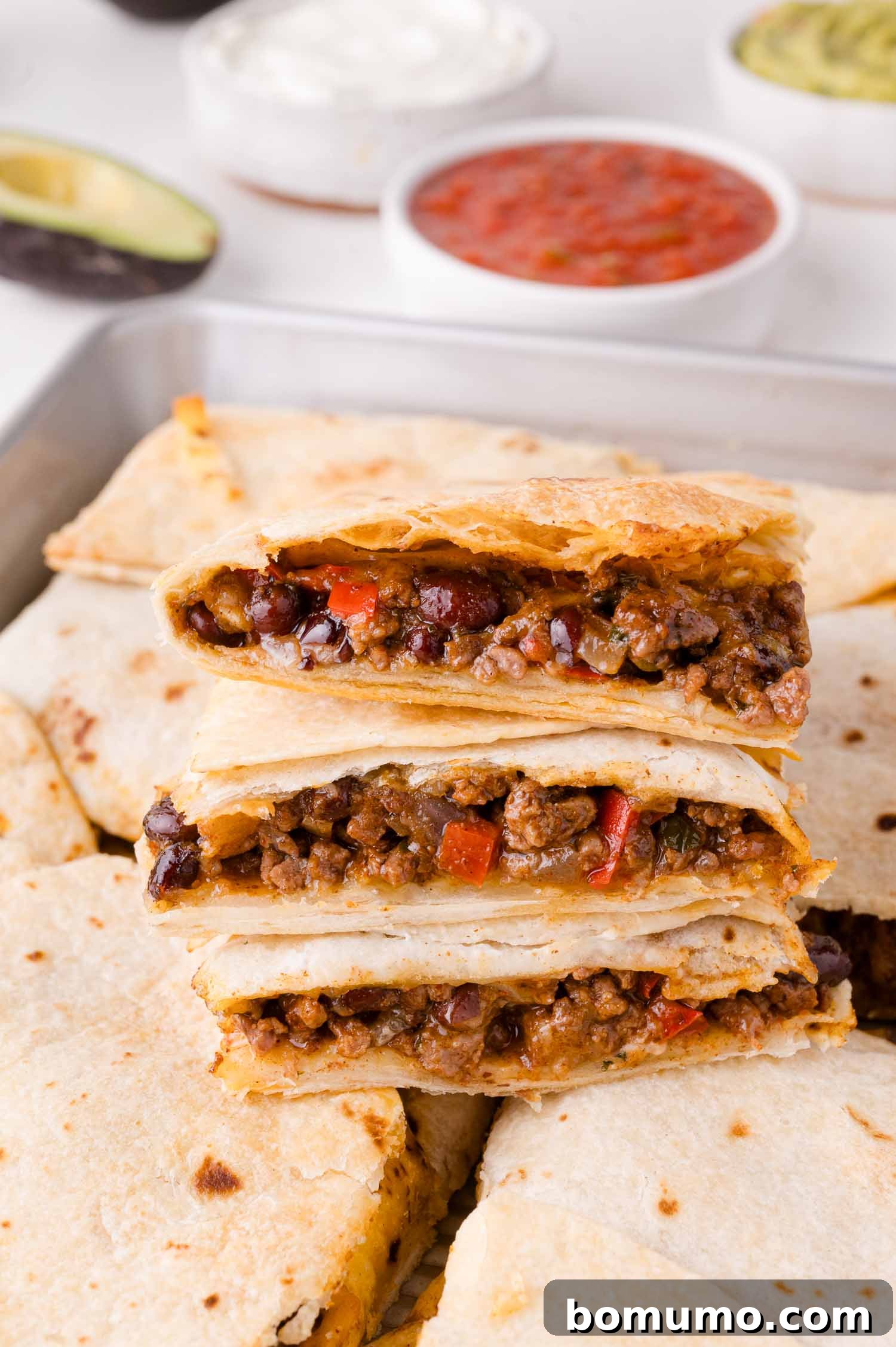 Effortless Sheet Pan Quesadillas 2 Three beef and bean quesadillas stacked on a tray, with bowls of salsa, sour cream, and guacamole in the background, showcasing a perfect family meal.