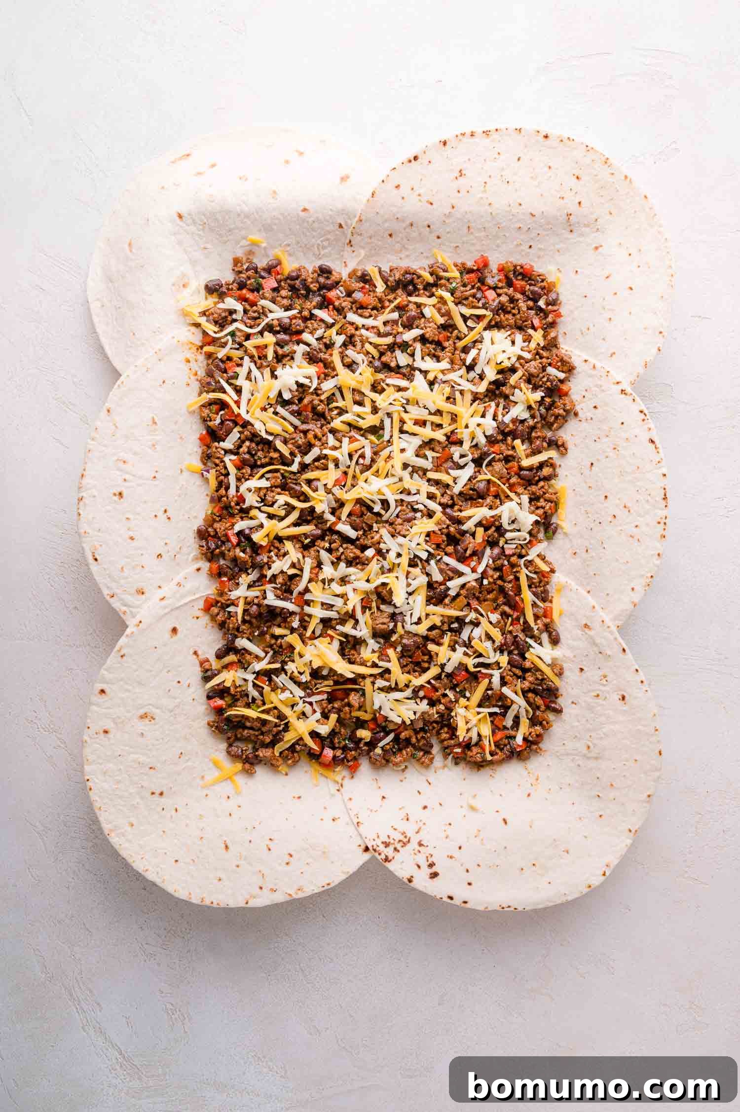 Effortless Sheet Pan Quesadillas 11 Six overlapping tortillas topped with cooked ground beef and grated cheese, placed on a light-colored surface, forming a cheesy, savory base.