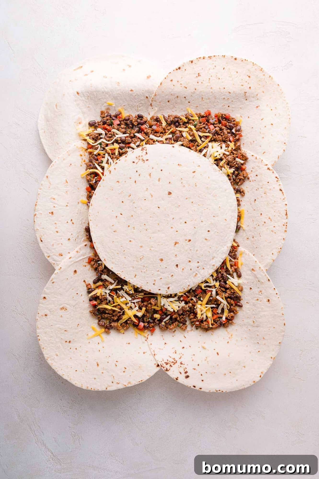 Effortless Sheet Pan Quesadillas 12 Six tortillas arranged on a sheet pan, filled with ground meat, diced vegetables, and shredded cheese, now covered and ready for baking.