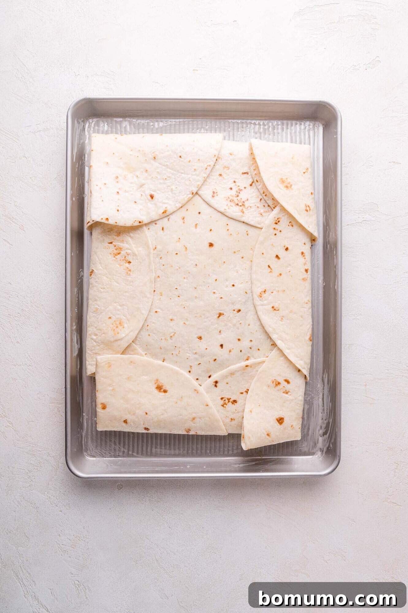 Effortless Sheet Pan Quesadillas 13 A metal baking sheet with overlapping tortillas arranged in a rectangular pattern, forming a sealed quesadilla.