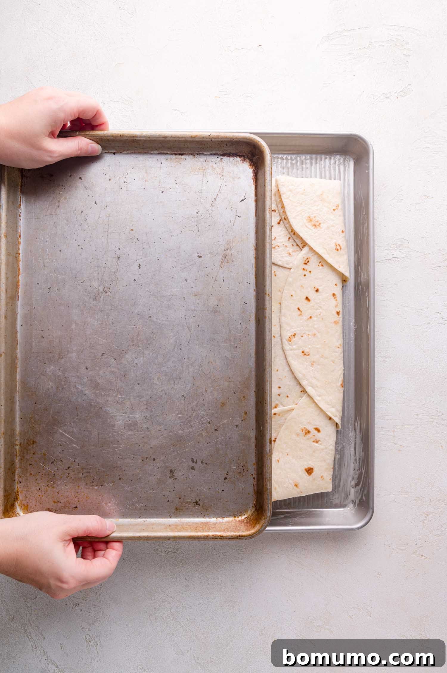 Effortless Sheet Pan Quesadillas 14 Hands carefully removing the top baking sheet from the sheet pan quesadillas, revealing their progress.