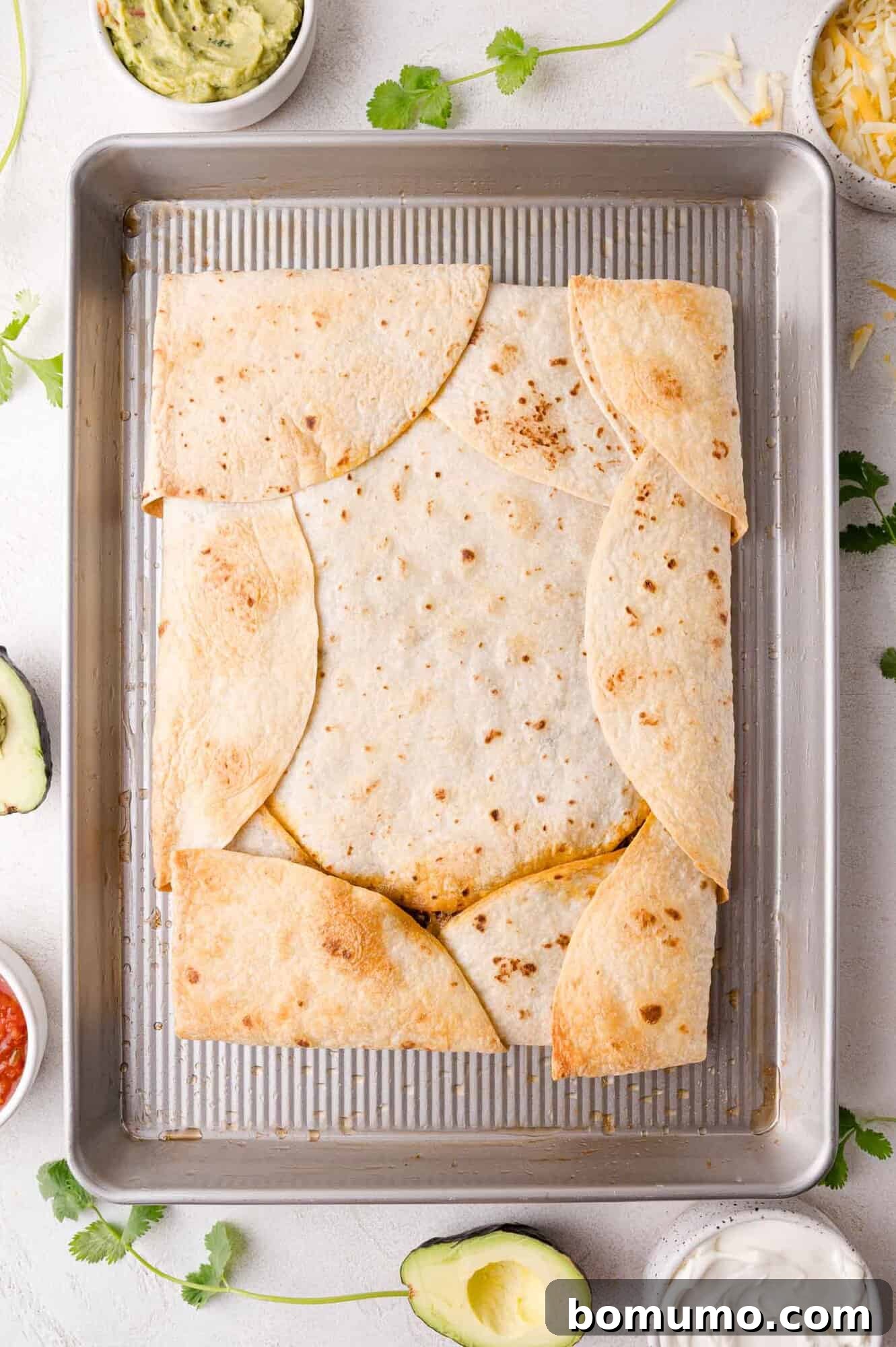 Effortless Sheet Pan Quesadillas 15 A baked sheet pan quesadilla on a tray, surrounded by ingredients like avocado, cilantro, salsa, and sour cream, showcasing its readiness.