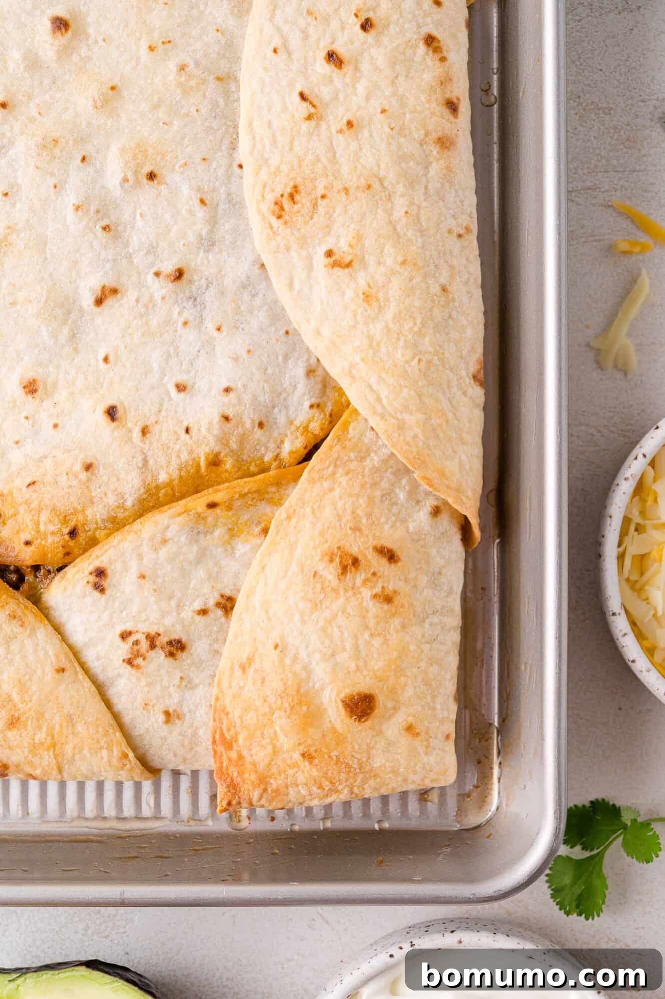 Effortless Sheet Pan Quesadillas 16 Baked quesadillas in a rectangular pan, cut into squares. Shredded cheese, avocado, cilantro, and sour cream are nearby for garnishing.