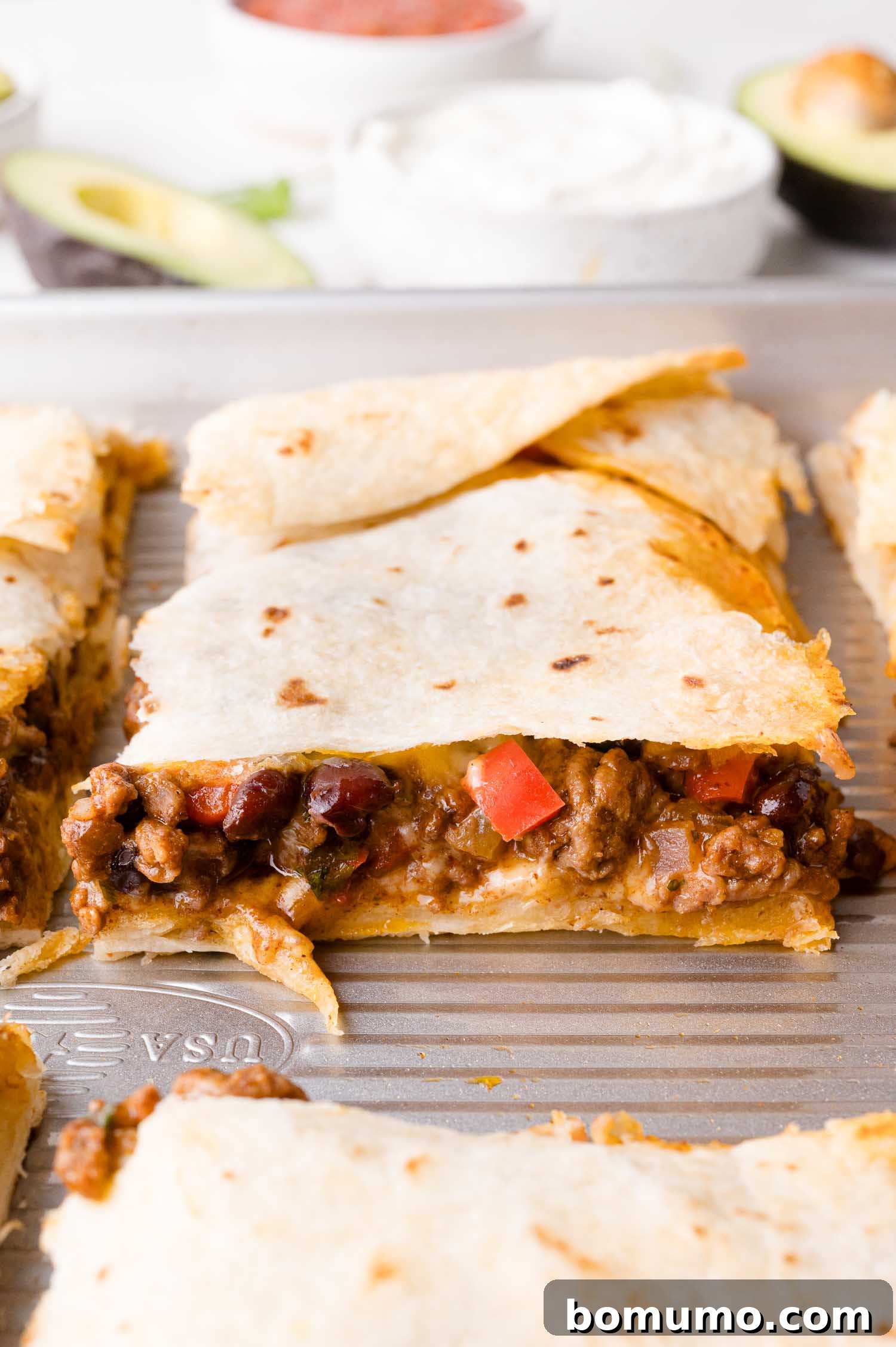 Effortless Sheet Pan Quesadillas 17 Close-up of a perfectly baked sheet pan quesadilla on a tray, filled with ground meat, black beans, and diced red bell peppers. Sliced avocado and bowls of sauce are visible in the background, inviting a delicious meal.