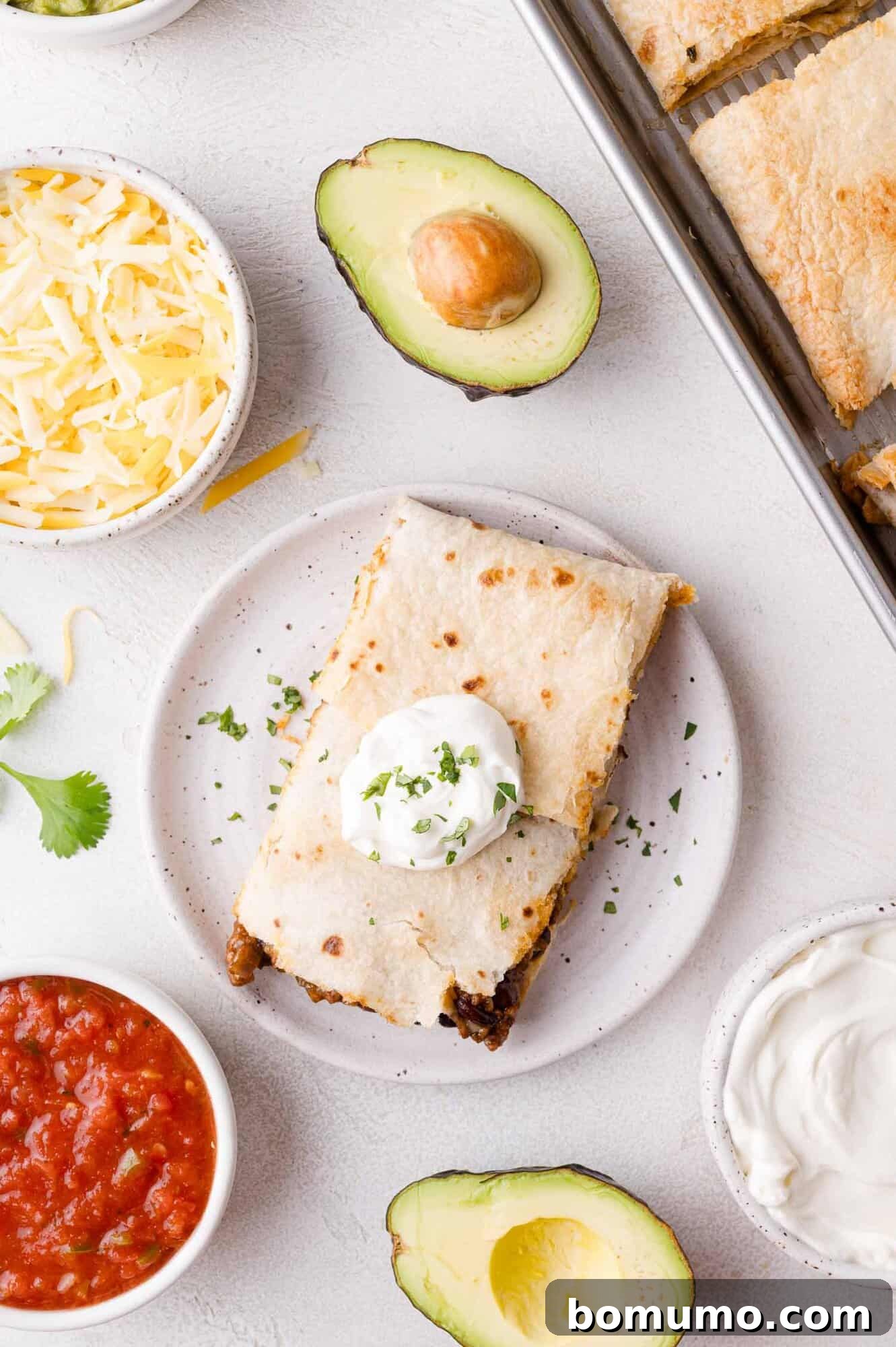 Effortless Sheet Pan Quesadillas 3 A plated sheet pan quesadilla topped with sour cream and herbs, surrounded by bowls of shredded cheese, salsa, sour cream, and avocado halves on a light surface, ready to be enjoyed.
