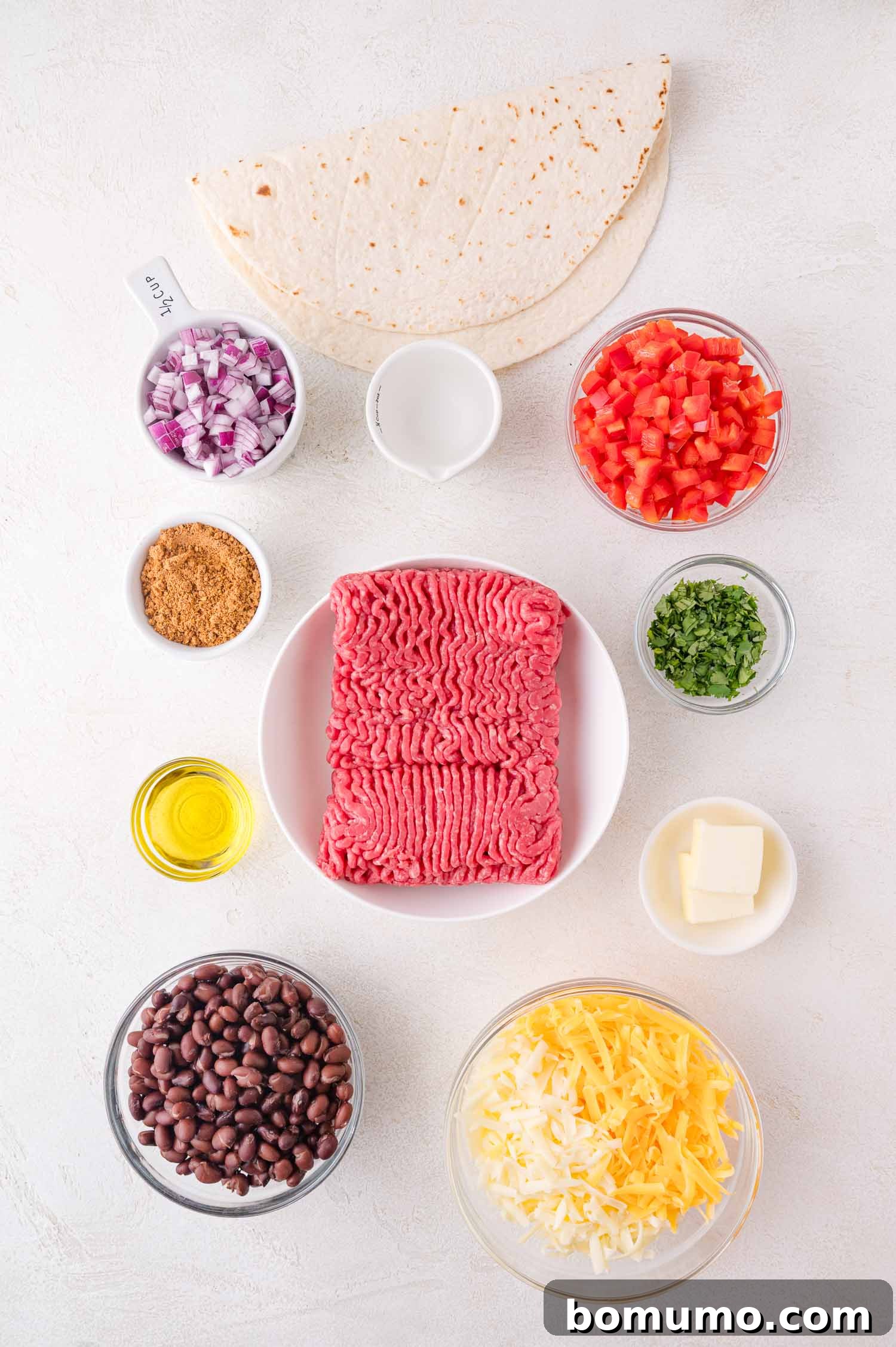 Effortless Sheet Pan Quesadillas 4 Ingredients for sheet pan quesadillas displayed: ground beef, tortillas, diced tomatoes, black beans, shredded cheese, chopped cilantro, diced onions, olive oil, sour cream, taco seasoning, and butter, all fresh and ready for cooking.