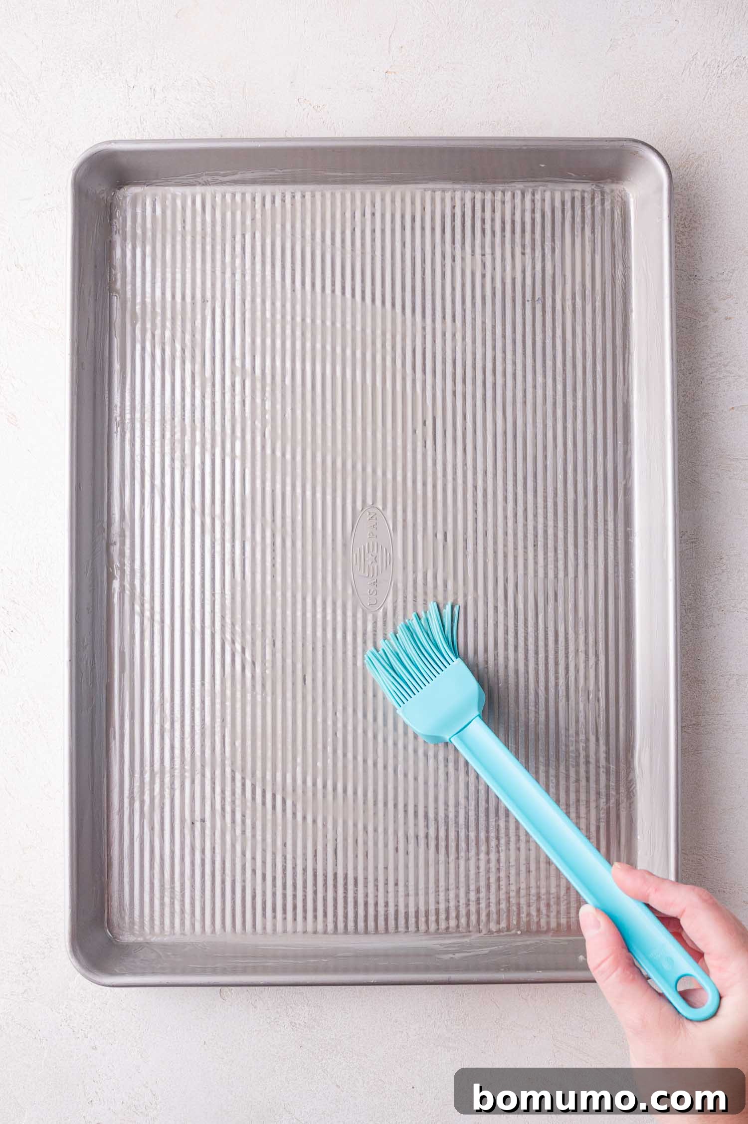 Effortless Sheet Pan Quesadillas 5 A hand uses a blue silicone brush to spread butter evenly on a ridged baking sheet, preparing it for the tortillas.