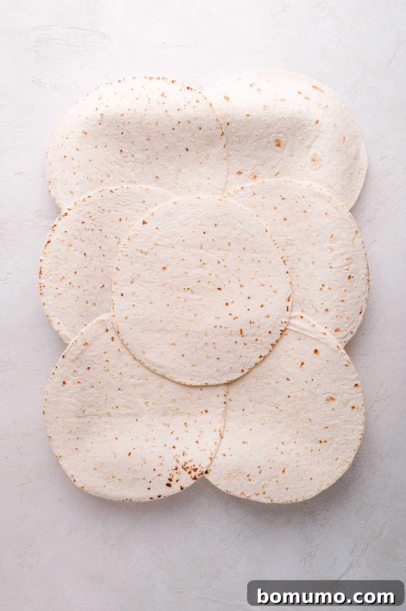 Effortless Sheet Pan Quesadillas 6 Seven flour tortillas are arranged in an overlapping, circular pattern on a light gray surface, covering the sheet pan's base.