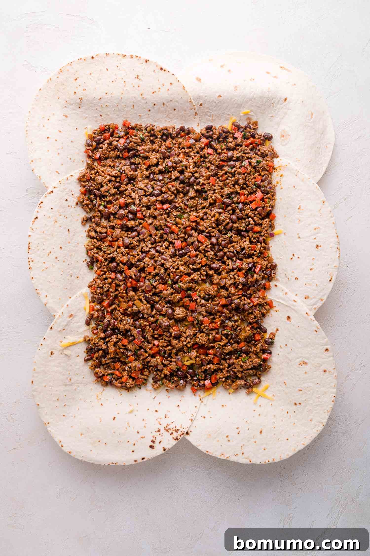Effortless Sheet Pan Quesadillas 10 Six tortillas arranged in a circle overlap slightly, topped with ground meat, black beans, and diced red peppers, spread in the center on a light surface, ready for the next layer.