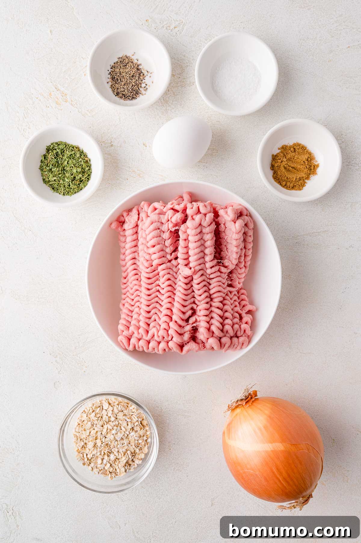 A beautifully composed shot showing a bowl of raw ground turkey, surrounded by smaller bowls containing various spices, quick oats, a whole egg, and an intact yellow onion, all arranged on a pristine white surface, ready for preparation.