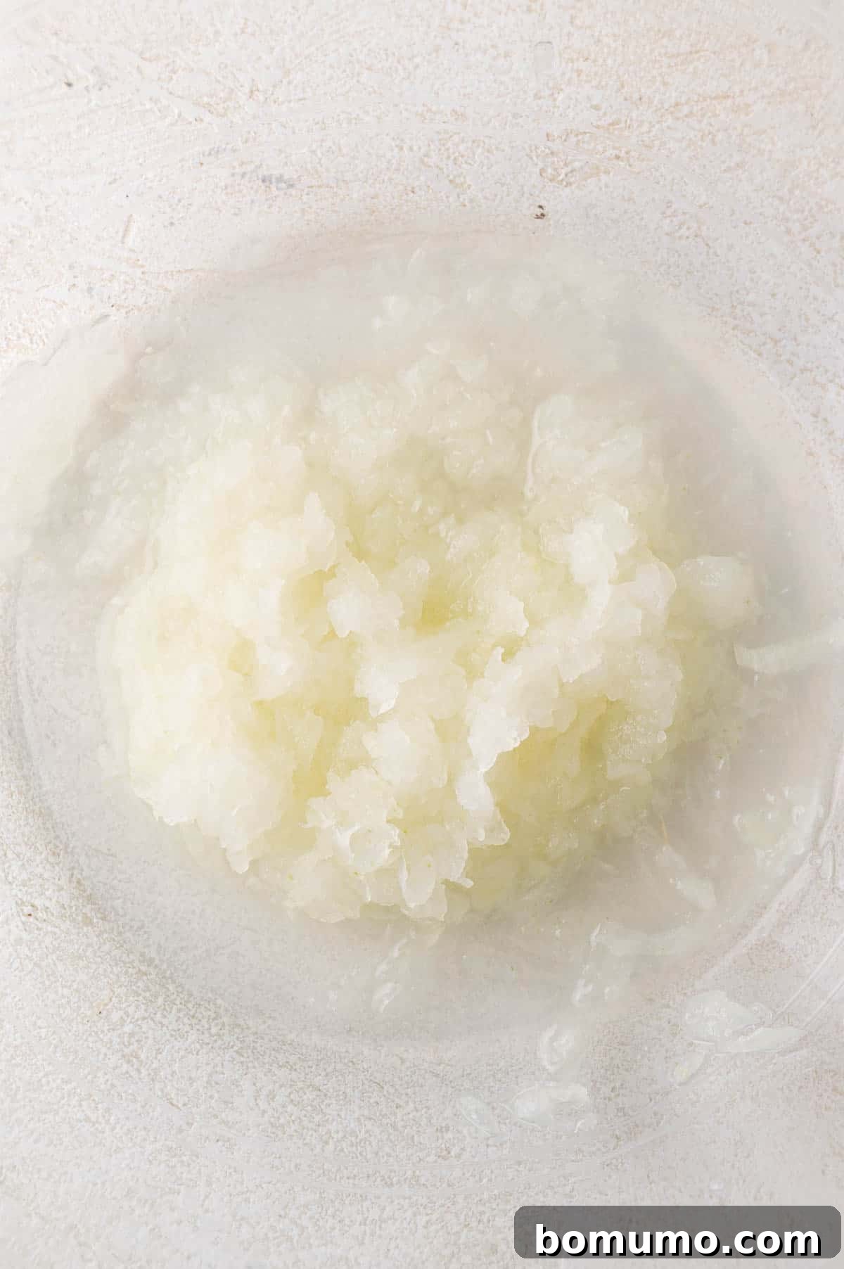 Finely grated yellow onion, along with its precious juices, collected neatly in a clear glass mixing bowl, set against a light-colored kitchen surface.