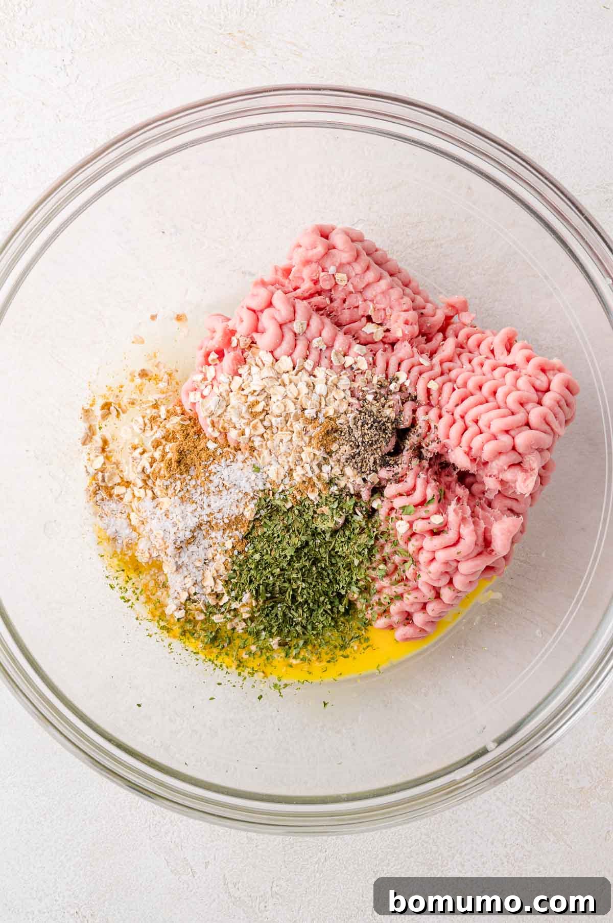 All the essential ingredients for turkey burgers – raw ground turkey, a beaten egg, quick oats, dried herbs, salt, pepper, and various spices – meticulously arranged in a large clear glass mixing bowl on a clean white surface, ready to be combined.