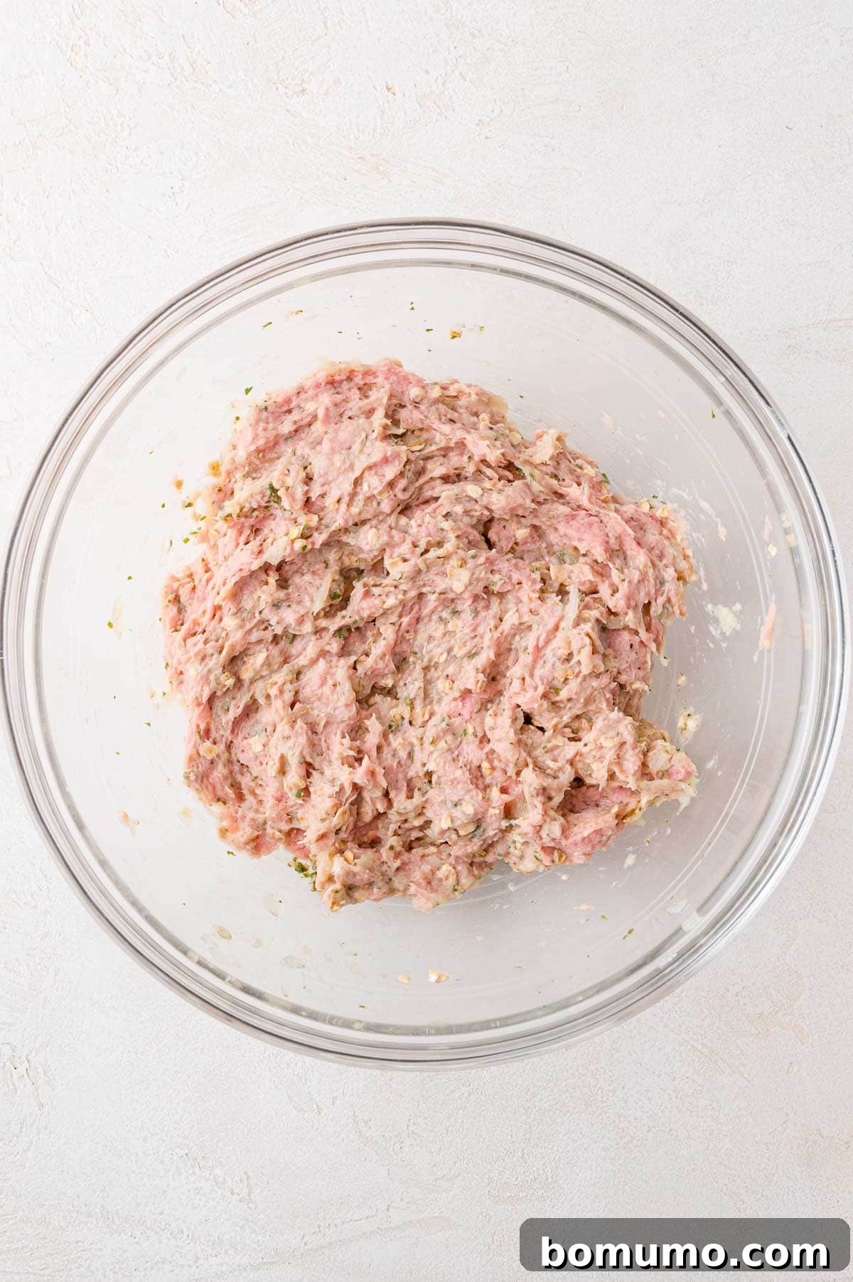 The fully mixed ground turkey, herb, and seasoning mixture is showcased in a clear glass bowl, ready for patty formation on a light-colored surface.