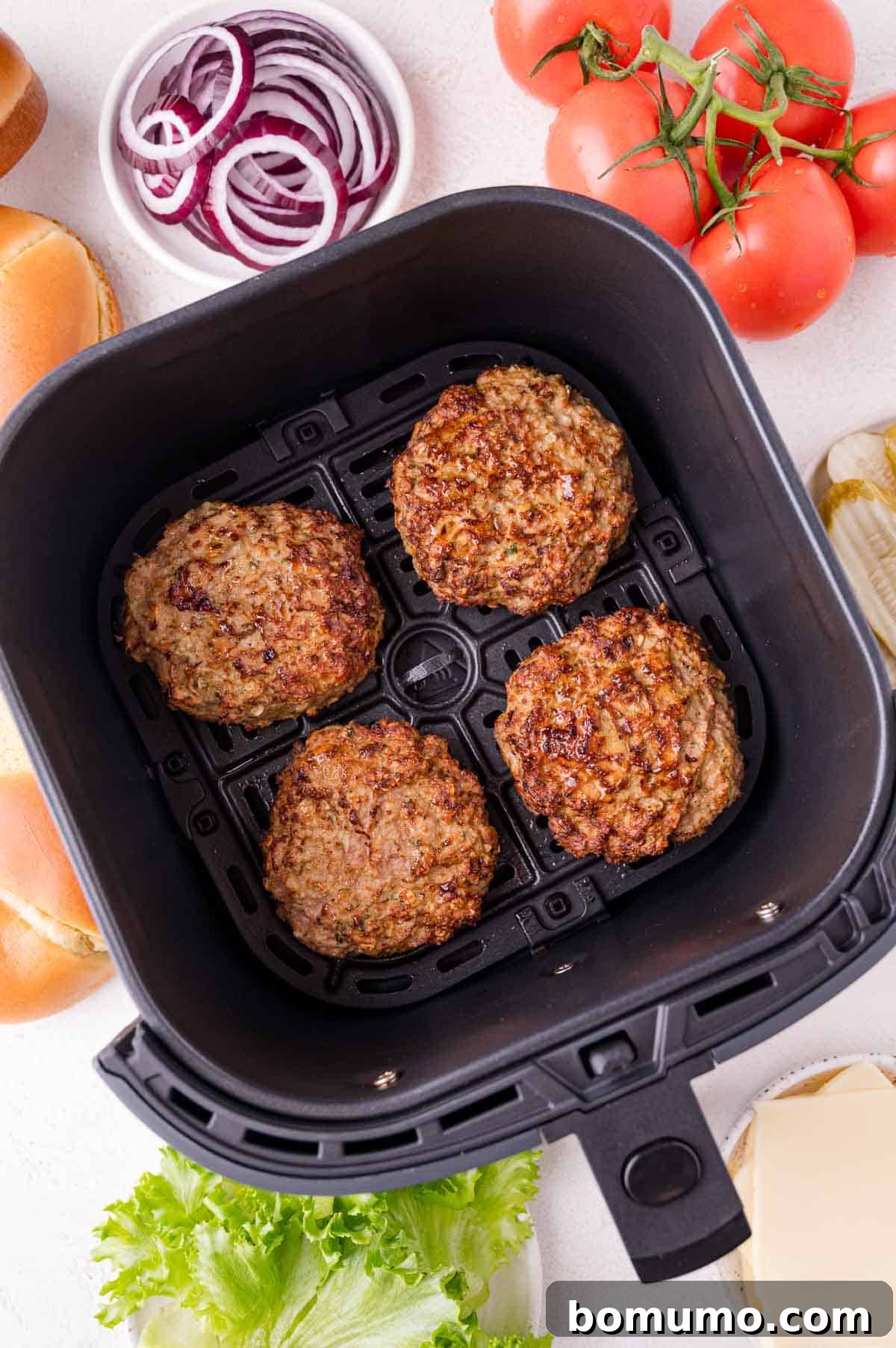 Four beautifully cooked burger patties, still in the air fryer basket, surrounded by an inviting spread of fresh ingredients including sliced red onions, whole ripe tomatoes, crisp lettuce, cheese slices, and soft burger buns, all arranged on a clean white surface.