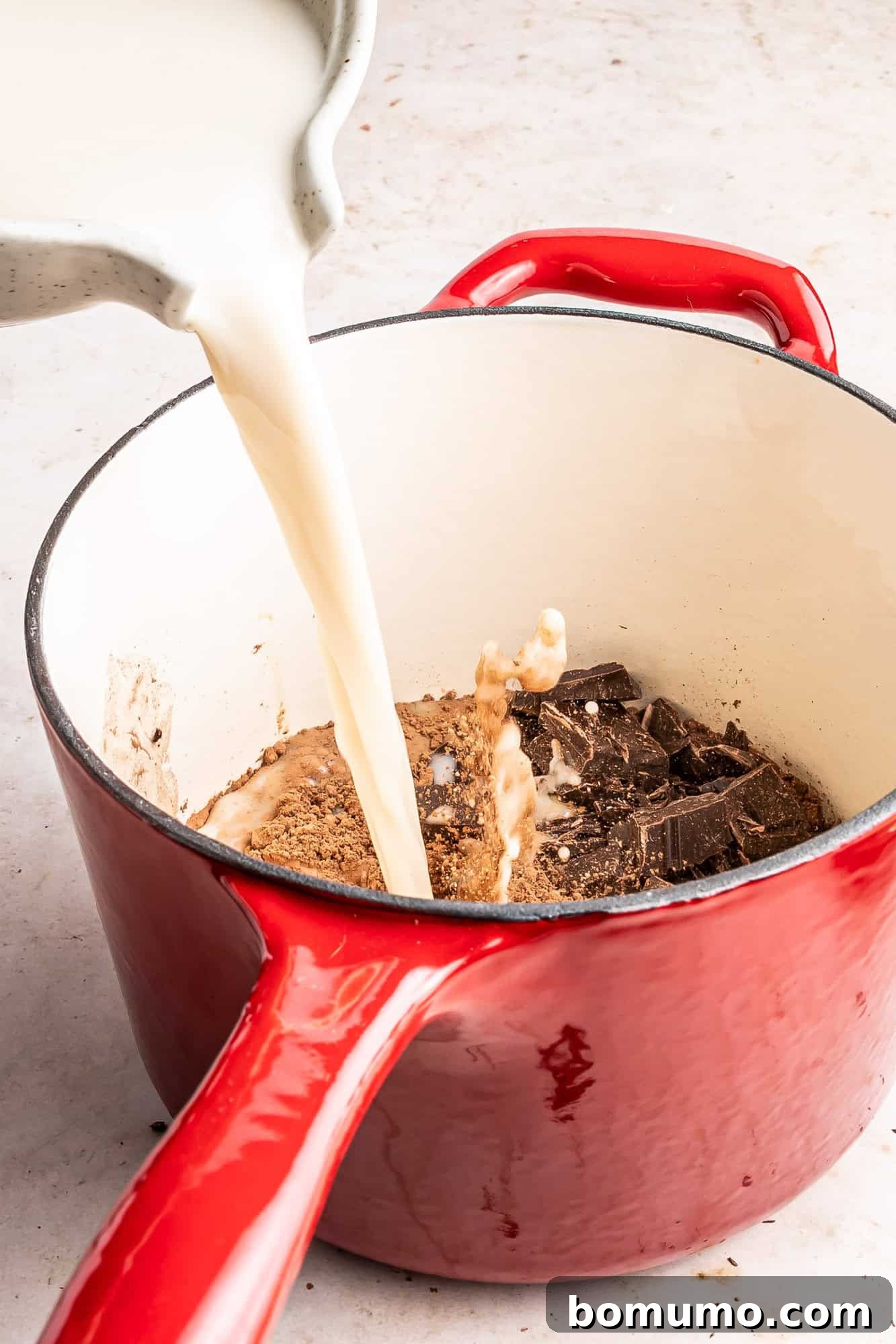Oat milk being poured into a small red saucepan containing dry ingredients, beginning the hot chocolate preparation.
