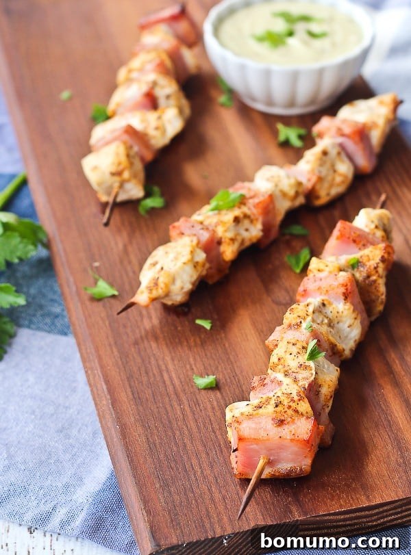 Overhead shot of three golden-brown chicken cordon bleu kabobs arranged on a rustic wooden cutting board, with a small white bowl of creamy Swiss cheese dipping sauce beside them.