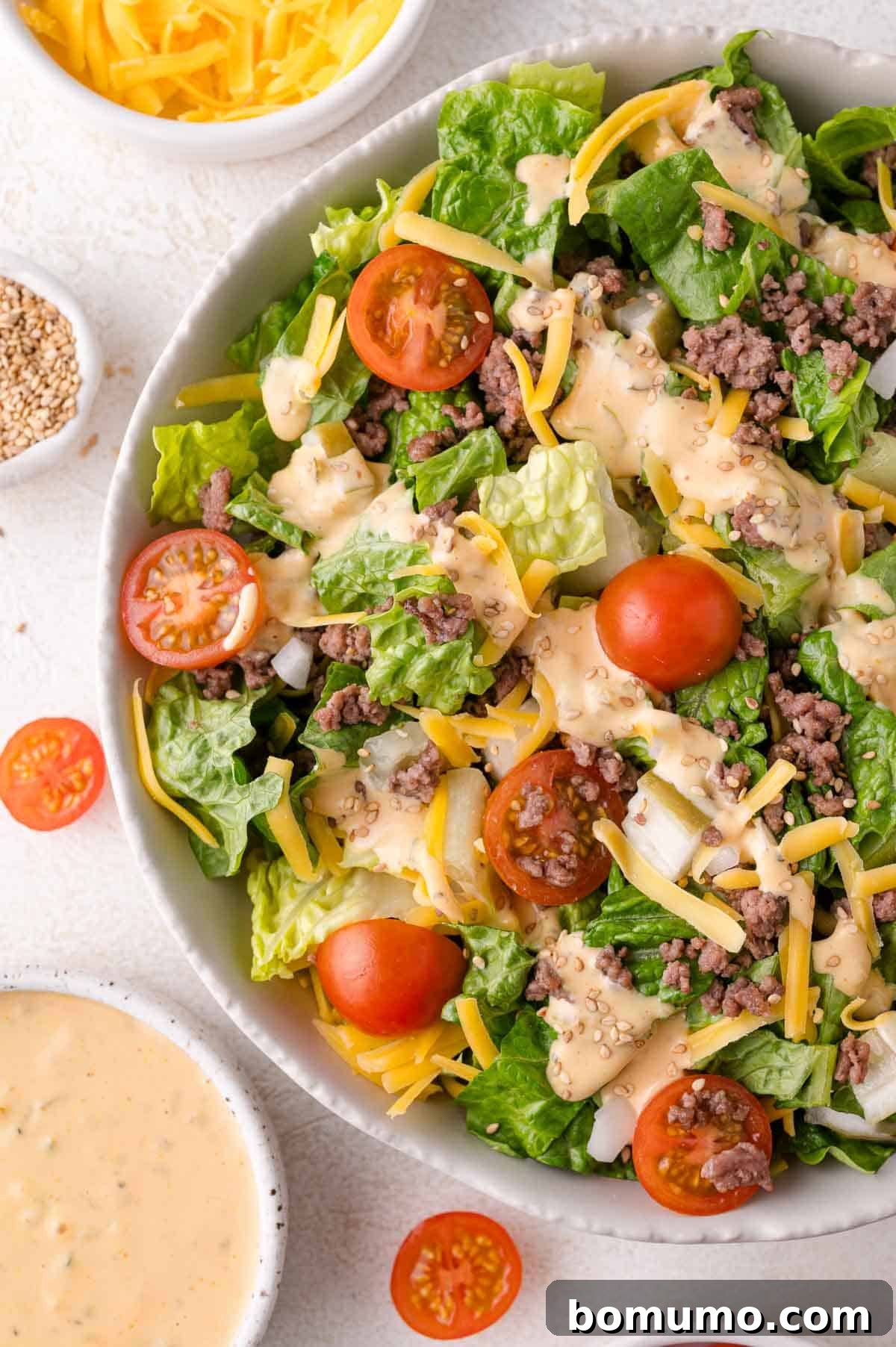 Classic Cheeseburger Salad 2 A vibrant Big Mac Salad featuring crisp romaine lettuce, seasoned ground beef, bright cherry tomatoes, shredded cheddar cheese, and a generous drizzle of creamy special sauce, garnished with sesame seeds. Small bowls of additional toppings like cheese, sesame seeds, and dressing are arranged nearby.