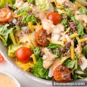 Classic Cheeseburger Salad 11 Salad with lettuce, cherry tomatoes, ground beef, shredded cheese, onions, and sesame seed topping in a white bowl.