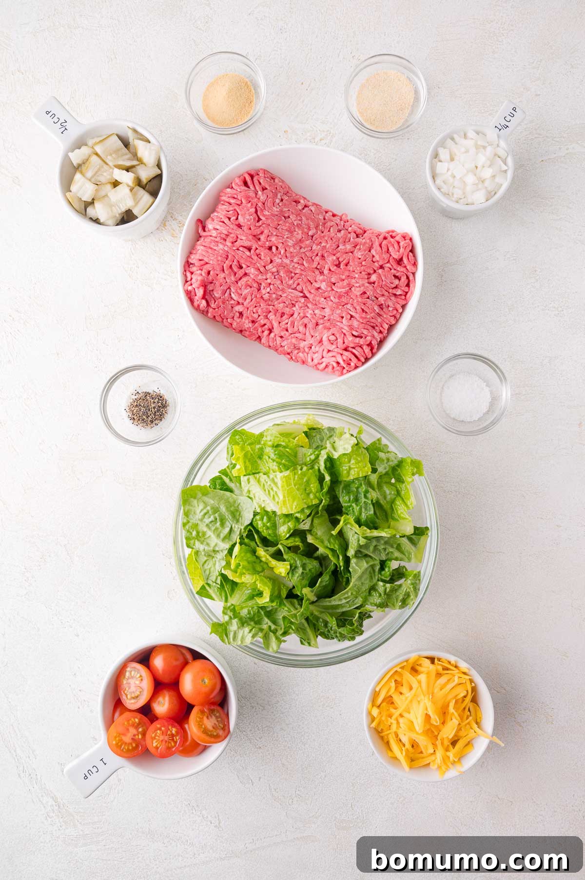 Classic Cheeseburger Salad 3 Ingredients for Big Mac Salad laid out on a table: lean ground beef, crisp lettuce, vibrant cherry tomatoes, rich cheddar cheese, aromatic diced onions, a selection of seasonings, and tangy dill pickles, all prepped and ready for assembly.