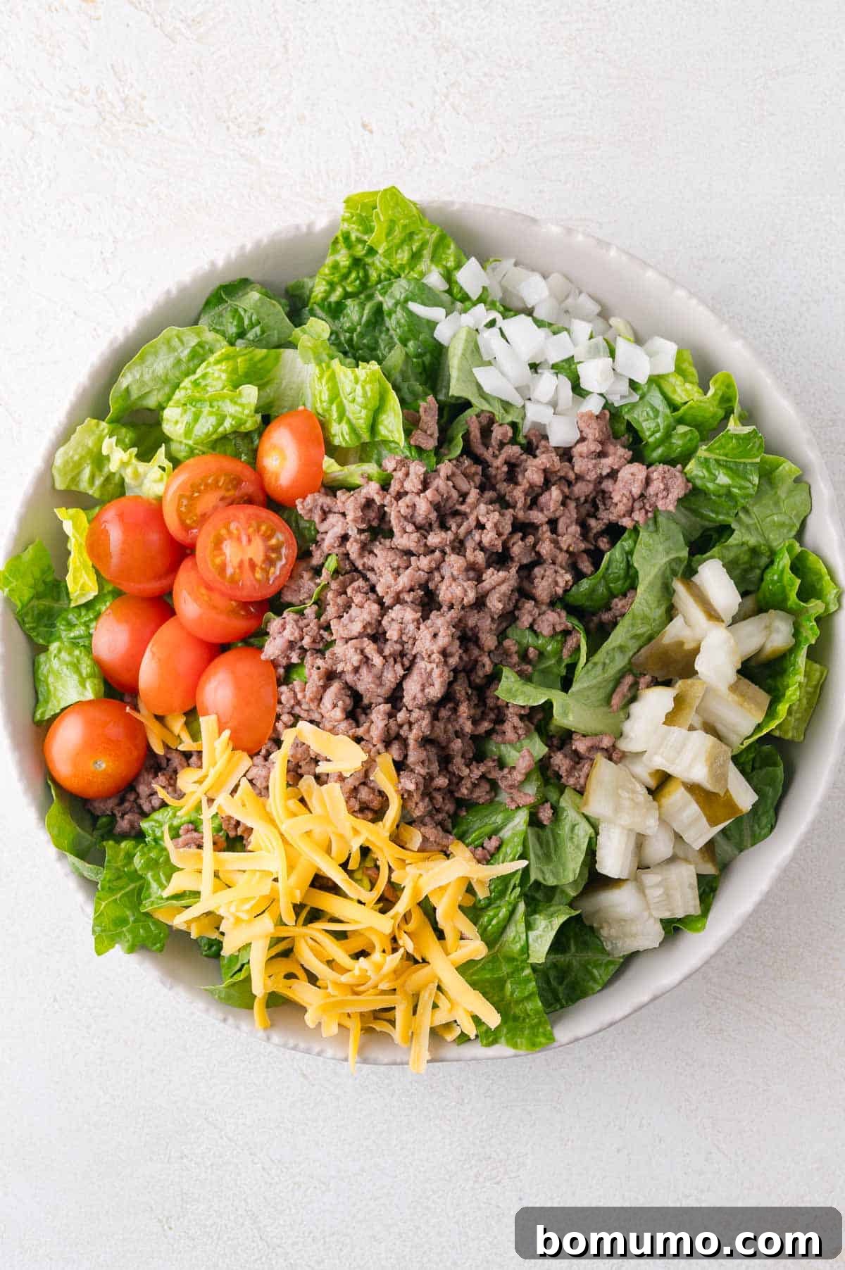 Classic Cheeseburger Salad 7 A vibrant Big Mac Salad in a bowl, showcasing distinct sections of fresh romaine lettuce, juicy halved cherry tomatoes, savory ground beef, shredded cheddar cheese, crisp diced onions, and tangy chopped pickles, artfully arranged for serving.