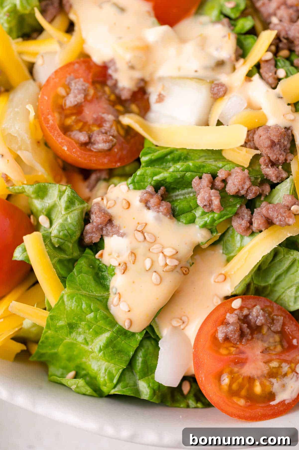 Classic Cheeseburger Salad 8 A close-up, appetizing view of a Big Mac Salad, featuring crisp romaine lettuce, tender ground meat, vibrant cherry tomatoes, melted shredded cheese, and a sprinkle of sesame seeds, all coated in the creamy, signature dressing.