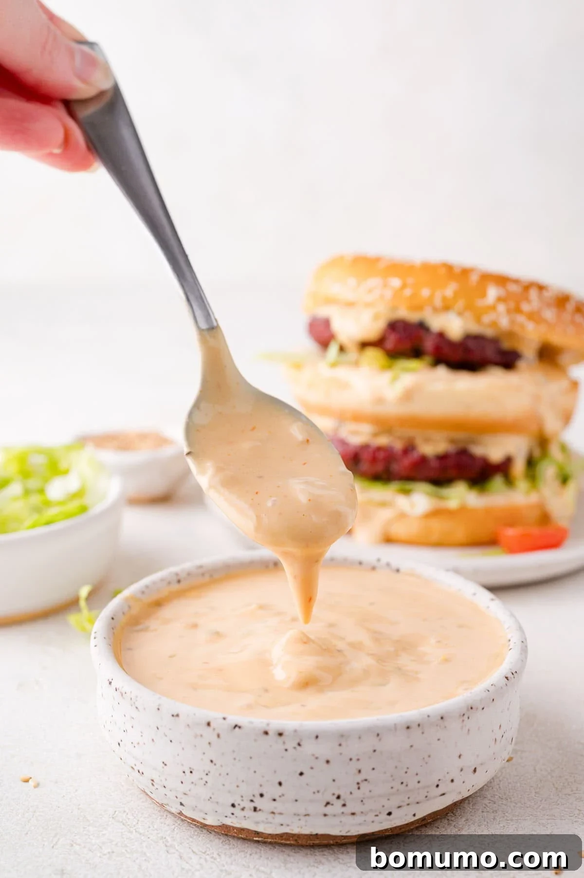 A spoon scoops creamy Big Mac sauce from a speckled bowl. Two burgers are stacked in the background alongside a bowl of shredded lettuce, hinting at the classic burger setup.