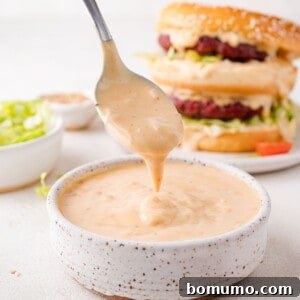 A spoon dripping creamy big mac sauce over a bowl. A hamburger with lettuce is in the background.