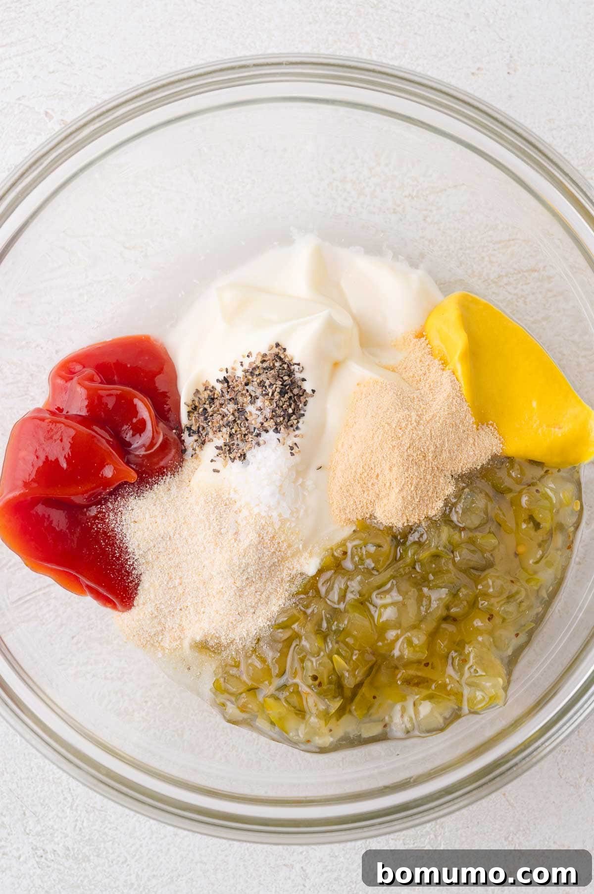Glass bowl containing ketchup, mayonnaise, mustard, pickle relish, garlic powder, onion powder, salt, pepper, and white powder on a light surface, showing all ingredients gathered before mixing.