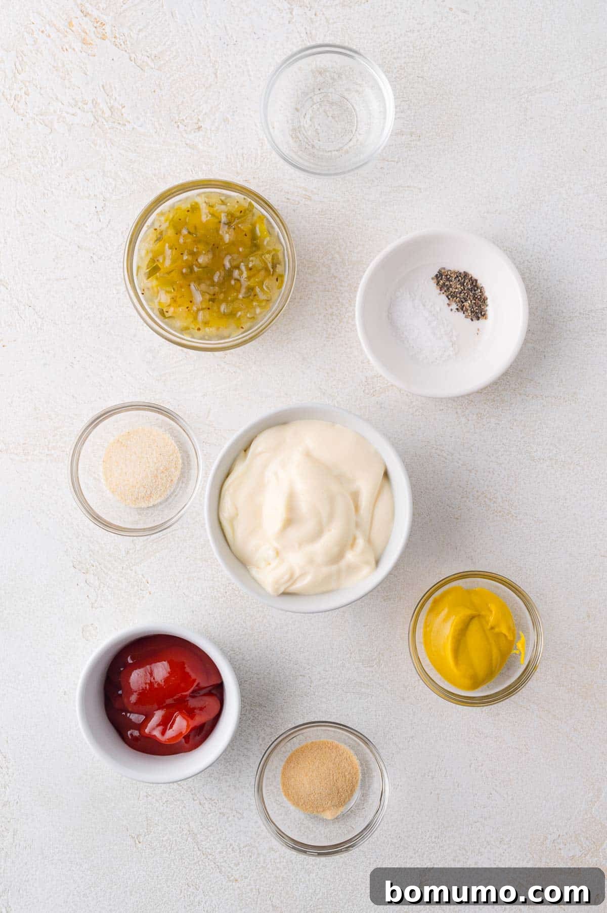 Ingredients for Big Mac sauce in small bowls on a white surface: mayonnaise, mustard, ketchup, relish, salt and pepper, garlic powder, and onion powder, showcasing neat preparation.