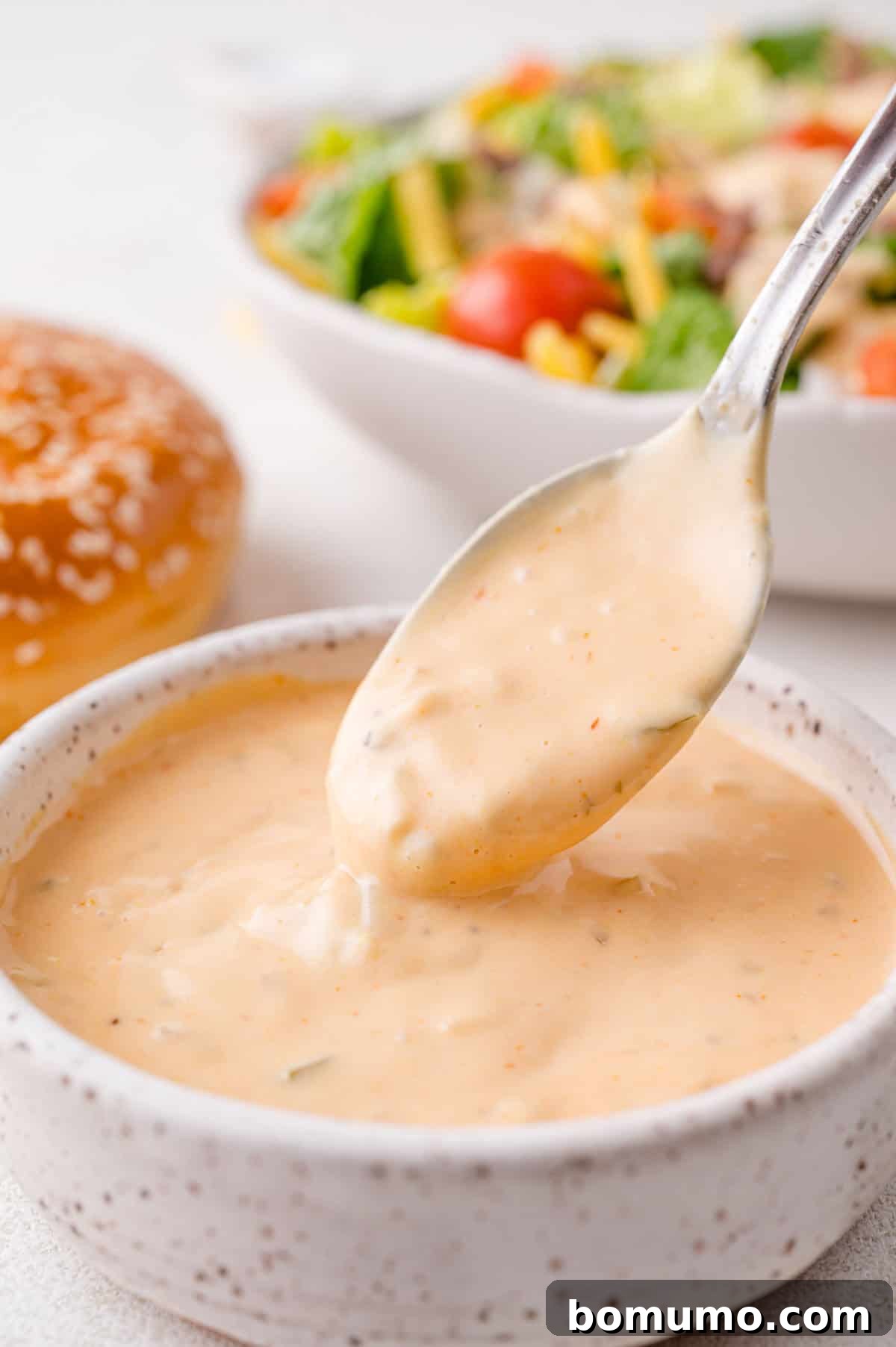 Spoon dipping into a bowl of creamy pink sauce, with a salad and a burger bun in the background, illustrating serving readiness.