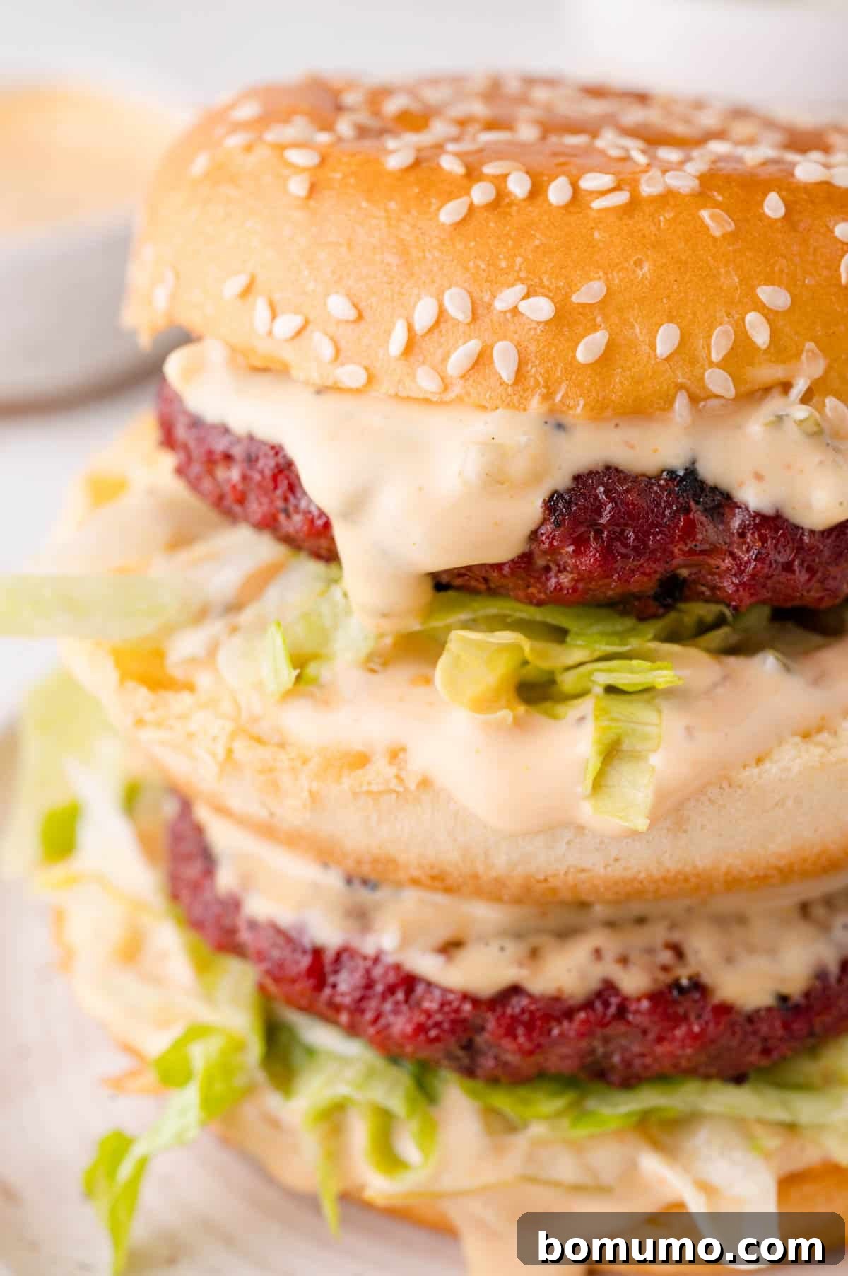Close-up of a double-decker burger with sesame seed bun, lettuce, and sauce, showcasing two patties and drizzled sauce for a mouth-watering presentation.