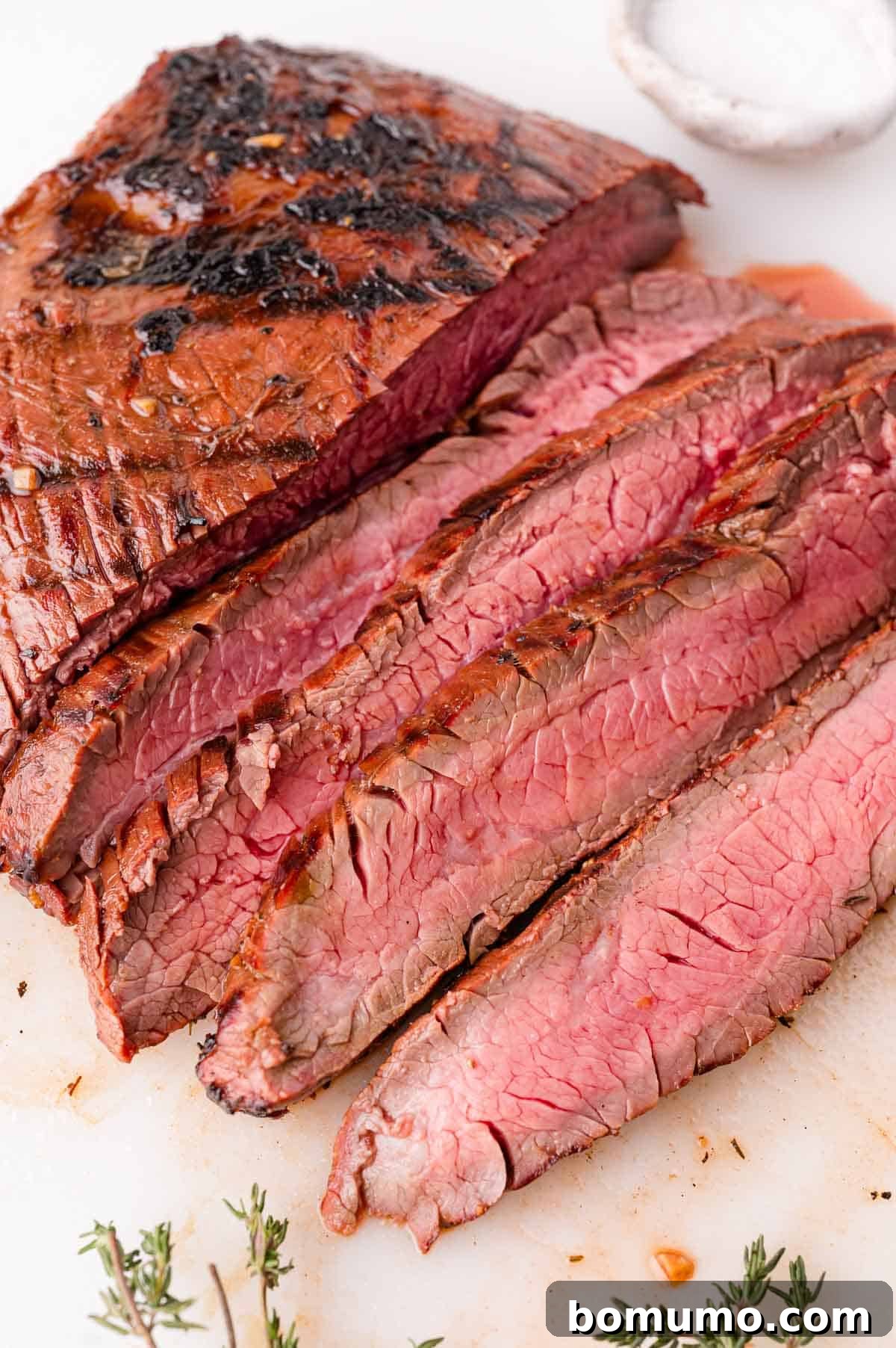 Sliced grilled steak on a cutting board with visible grill marks, served medium rare, with a small dish of salt and sprigs of fresh herbs nearby.
