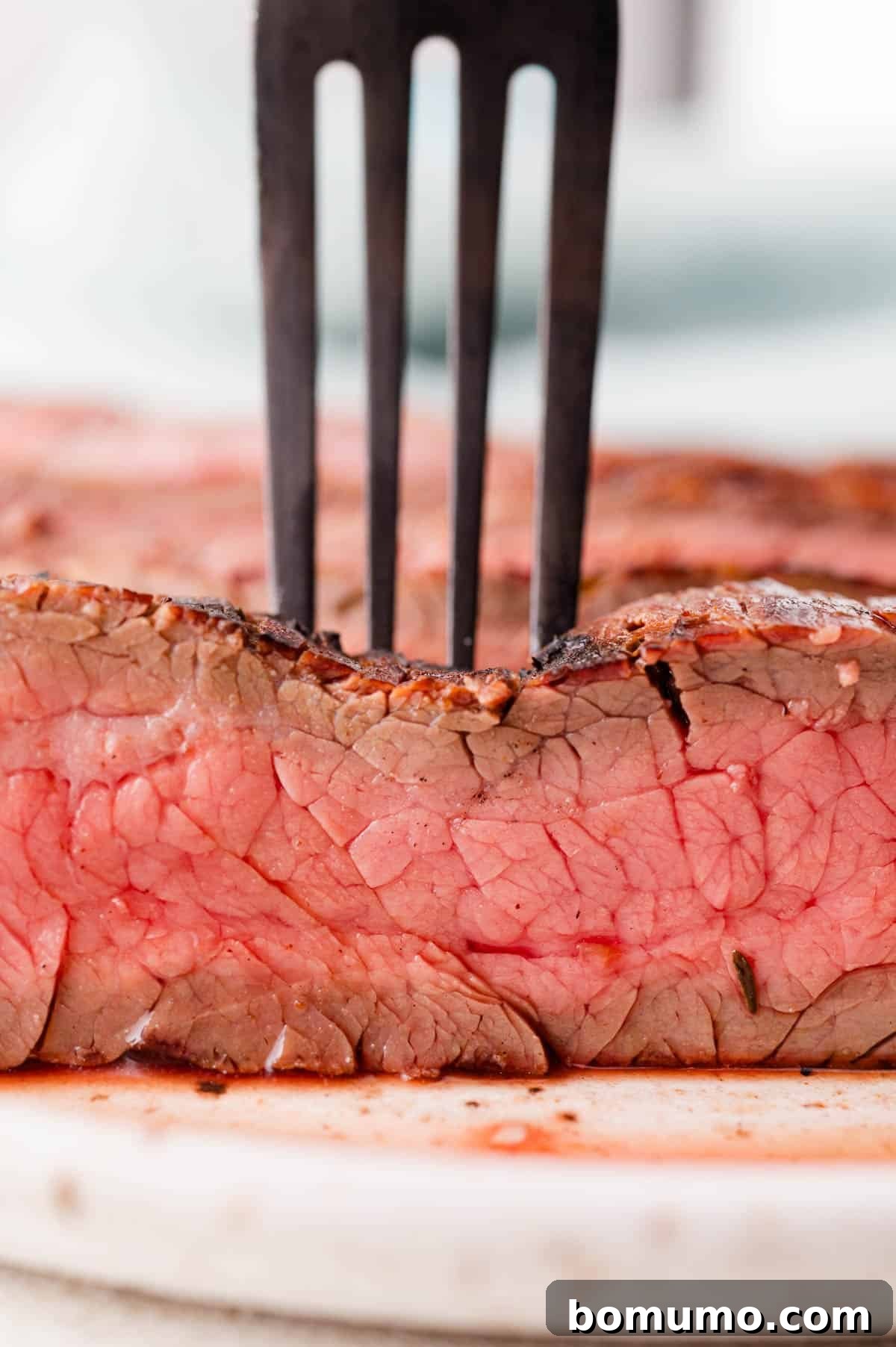 A metal fork pierces a thick slice of rare steak with a seared crust, showcasing its juicy pink interior on a white cutting board.